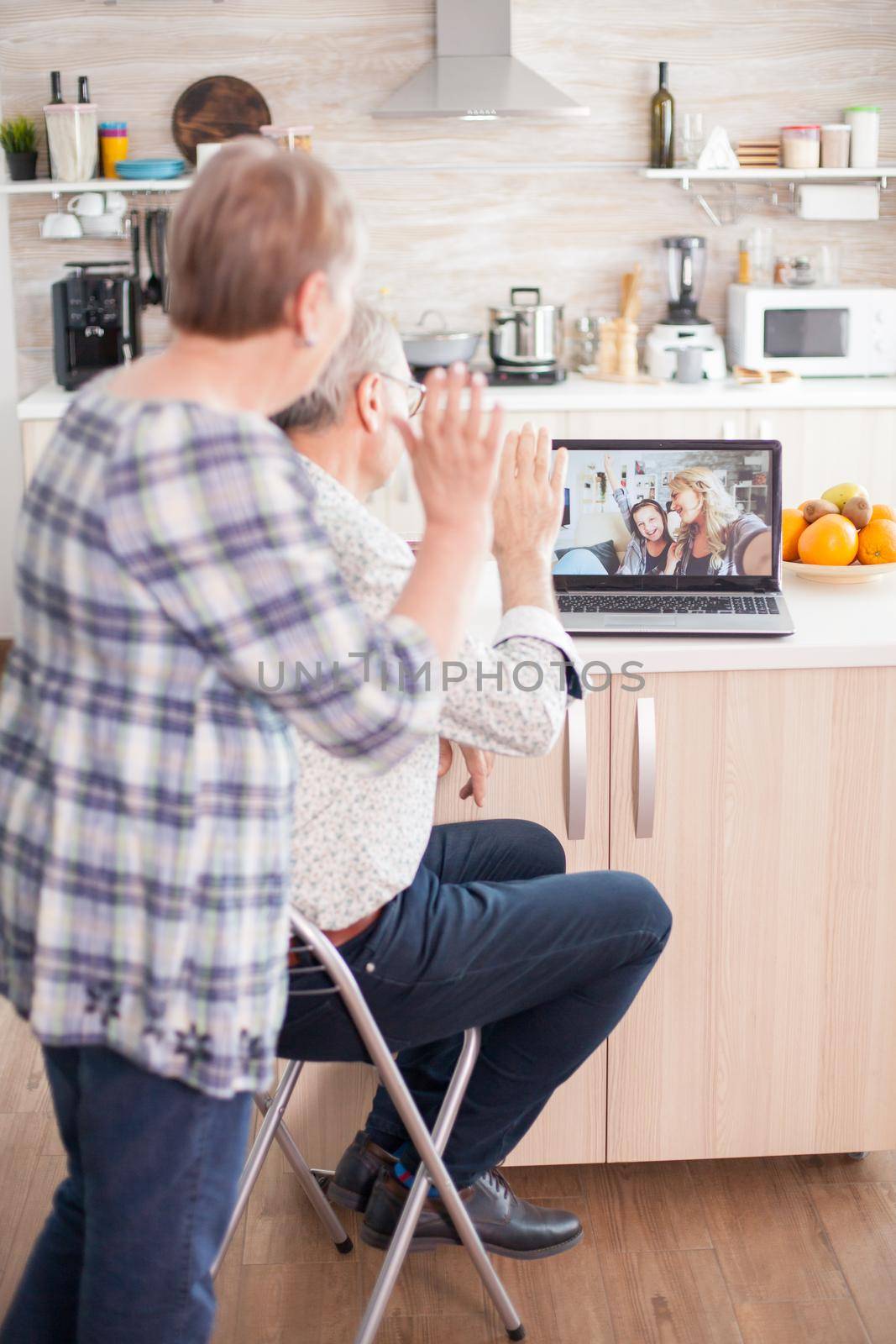 Senior couple saying hello to daughter and niece. Happy senior woman during a video conference with family using laptop in kitchen. Online call with daughter and niece. Elderly person using modern communication online internet web techonolgy.