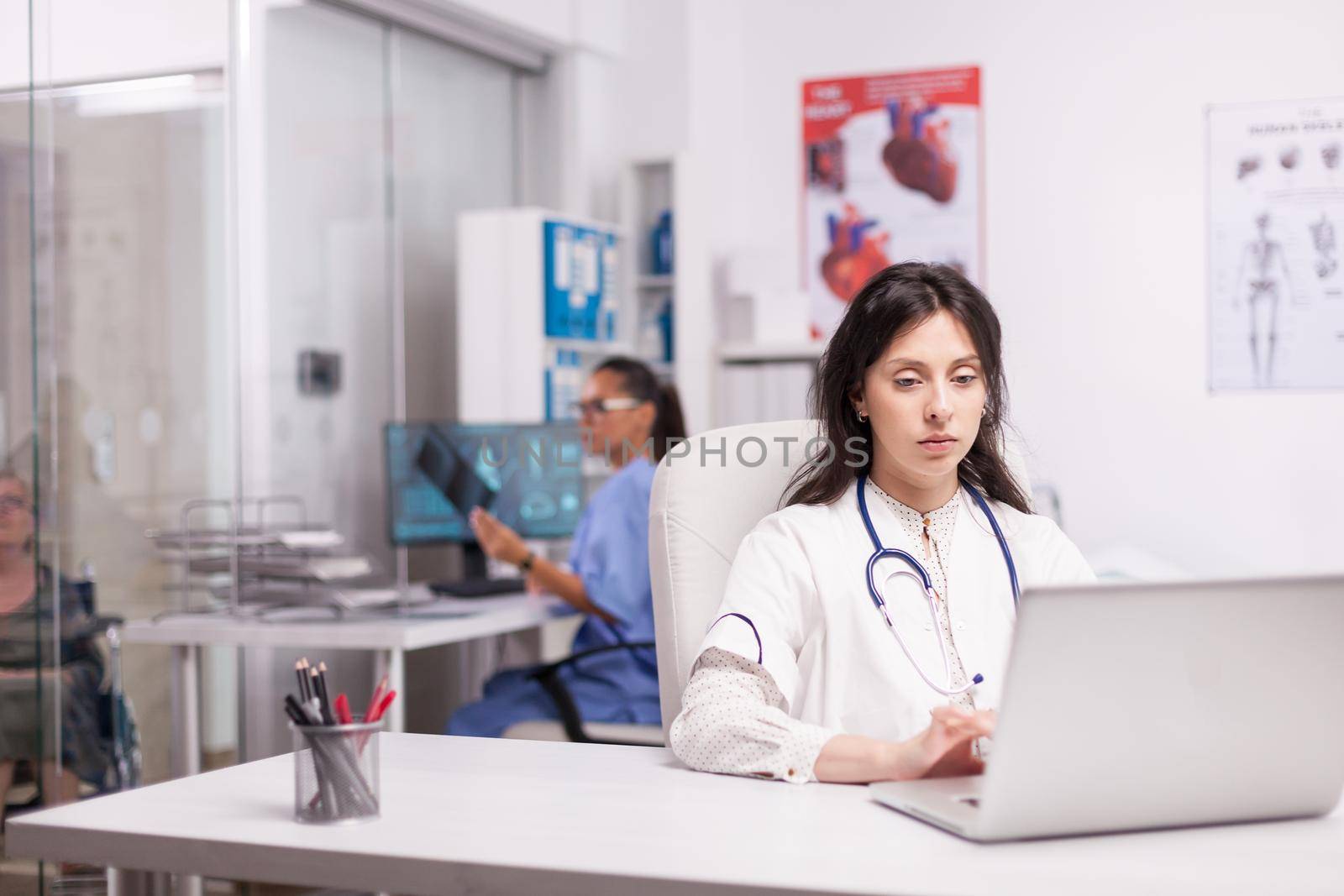 Young therapist using laptop in clinic office typing document wearing white coat with stethoscope and nurse is holding x-ray image while disabled senior patient in wheelchair is waiting.