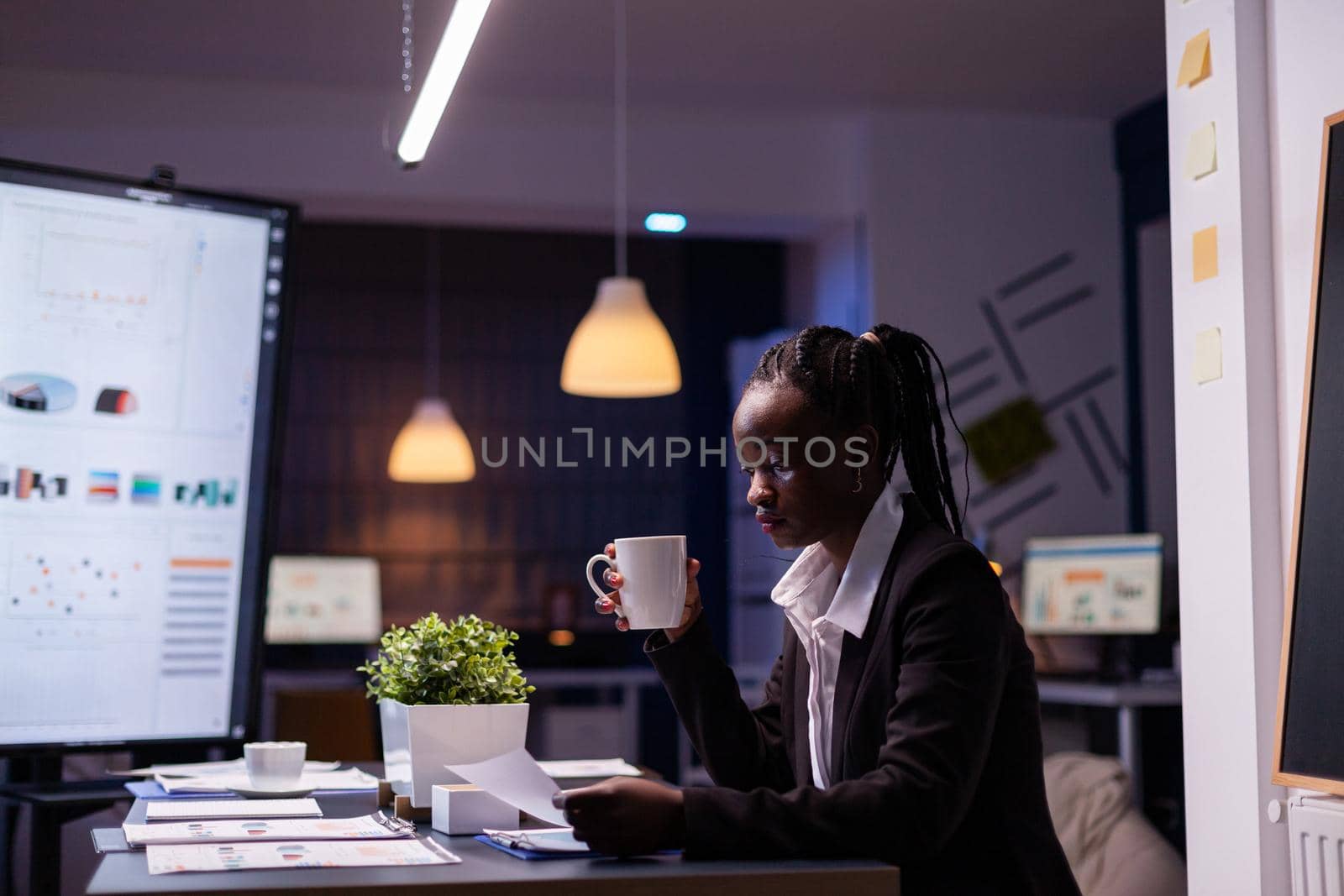 Workaholic african american businesswoman analysing marketing statistics on paperwork drinking coffee sitting at table late at night. Business manager with dark skin working in office meeting room