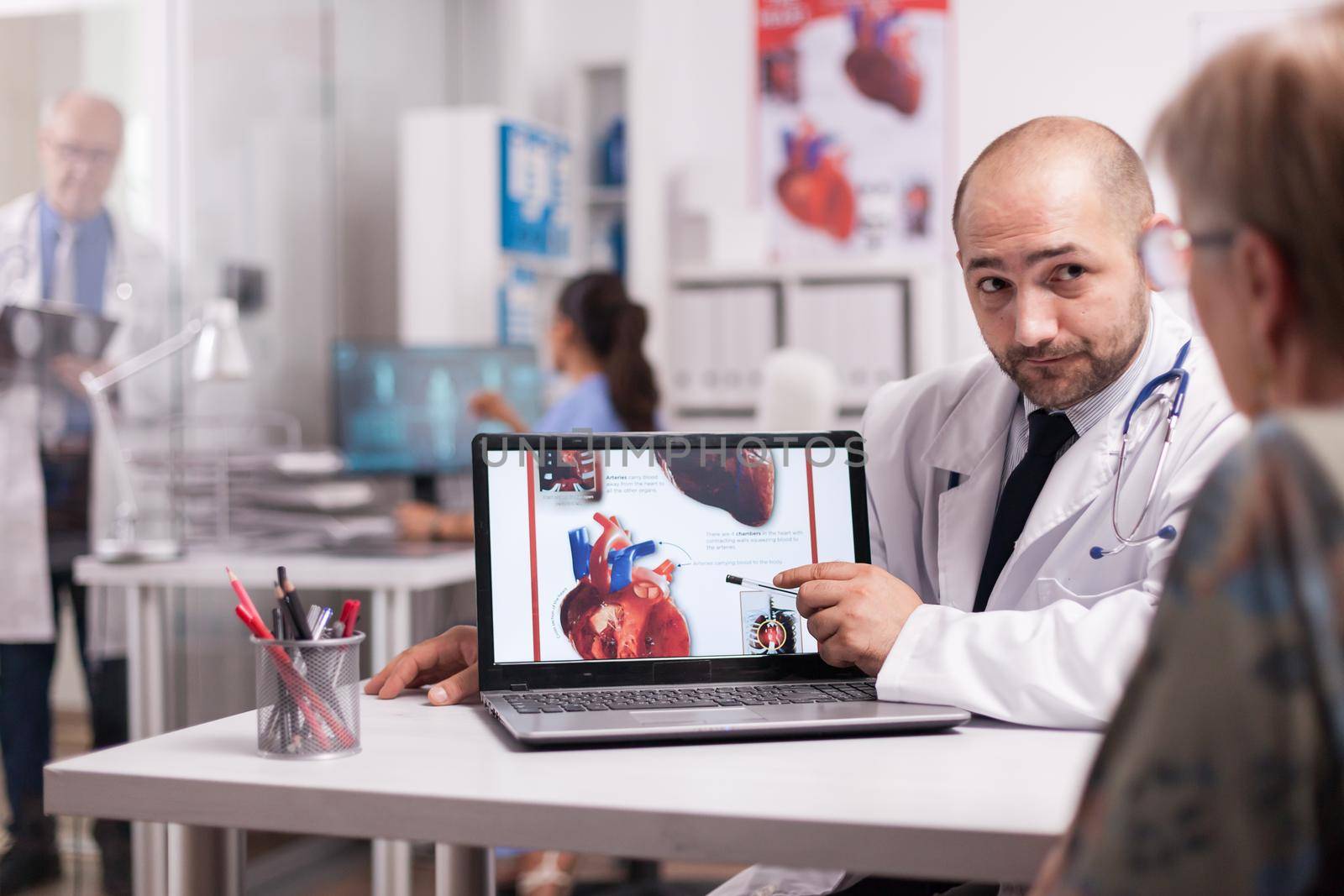 Doctor pointing at heart with trauma after senior patient heart attack. Elderly aged woman discussing about medication with young medic in hospital office. Mature medic wearing white coat and taking notes on clipboard.