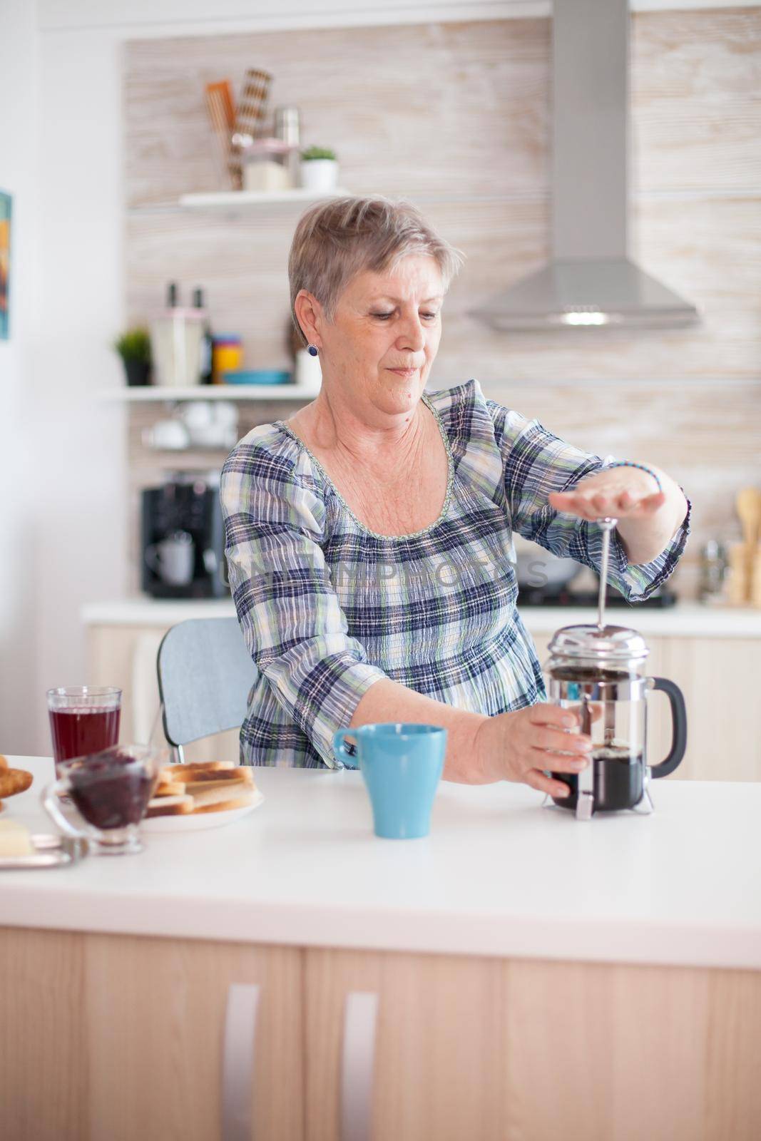 Senior woman pressing on french press to make tasty coffee during breakfast in kitchen. Elderly person in the morning enjoying fresh brown cafe espresso cup caffeine from vintage mug, filter relax refreshment