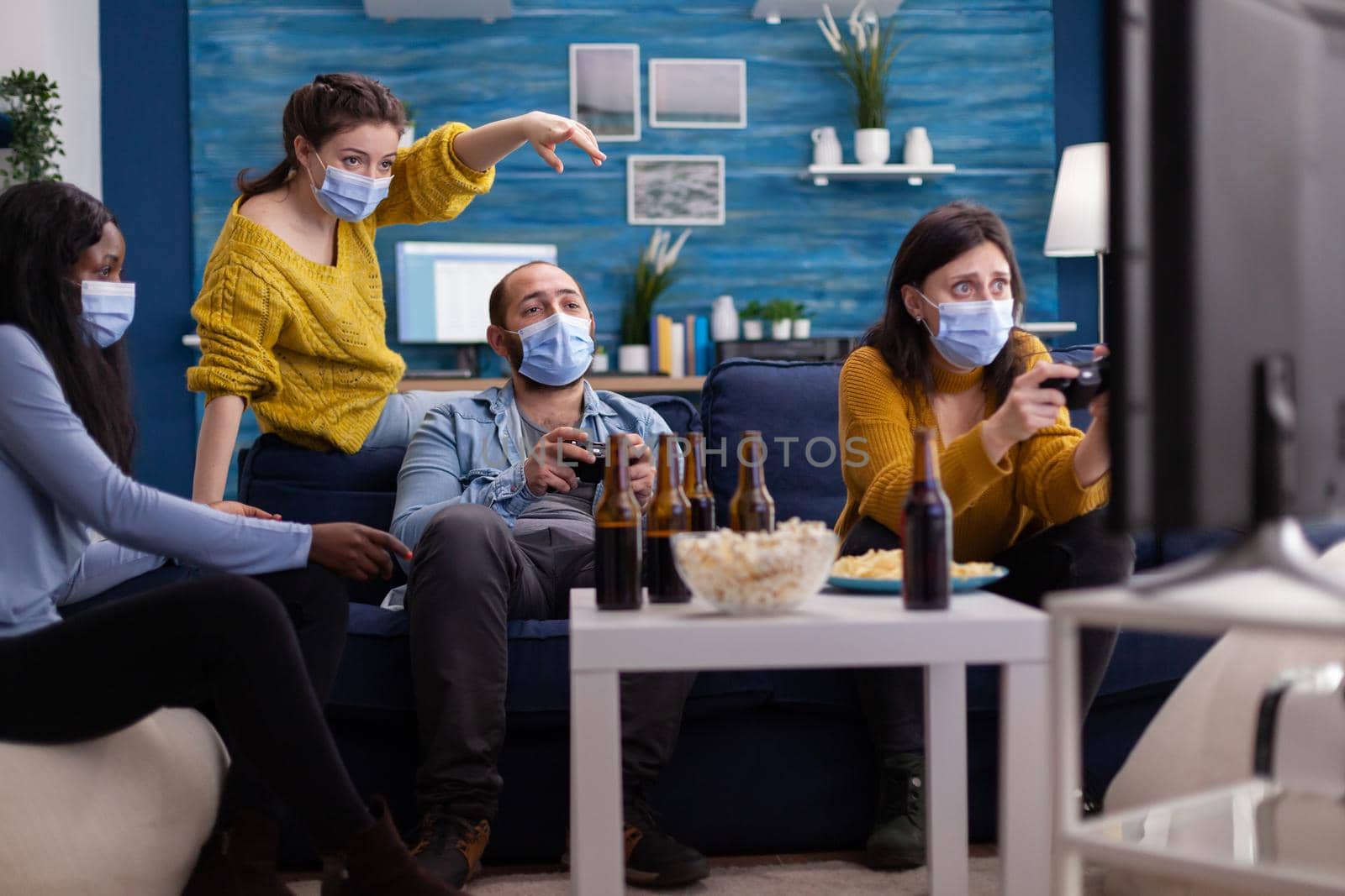 Cheerful multiracial friends playing video games with joystick controller in apartment living room wearing face mask to prevent spreading coronavirus. Conceptual image.
