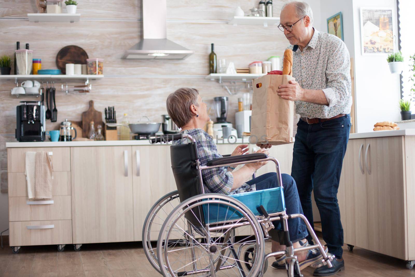 Senior man taking grocery paper bag from handicapped wifein wheelchair. Mature people with fresh vegetables from market. Living with disabled person with walking disabilities