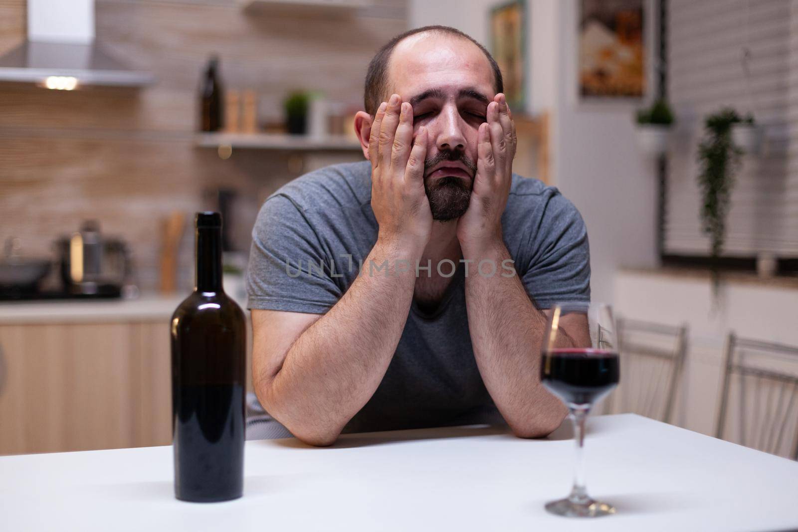 Desperate man feeling miserable while having glass with wine and bottle drinking alone. Person with unhealthy addiction sitting with liquor, booze, alcoholic beverage being upset