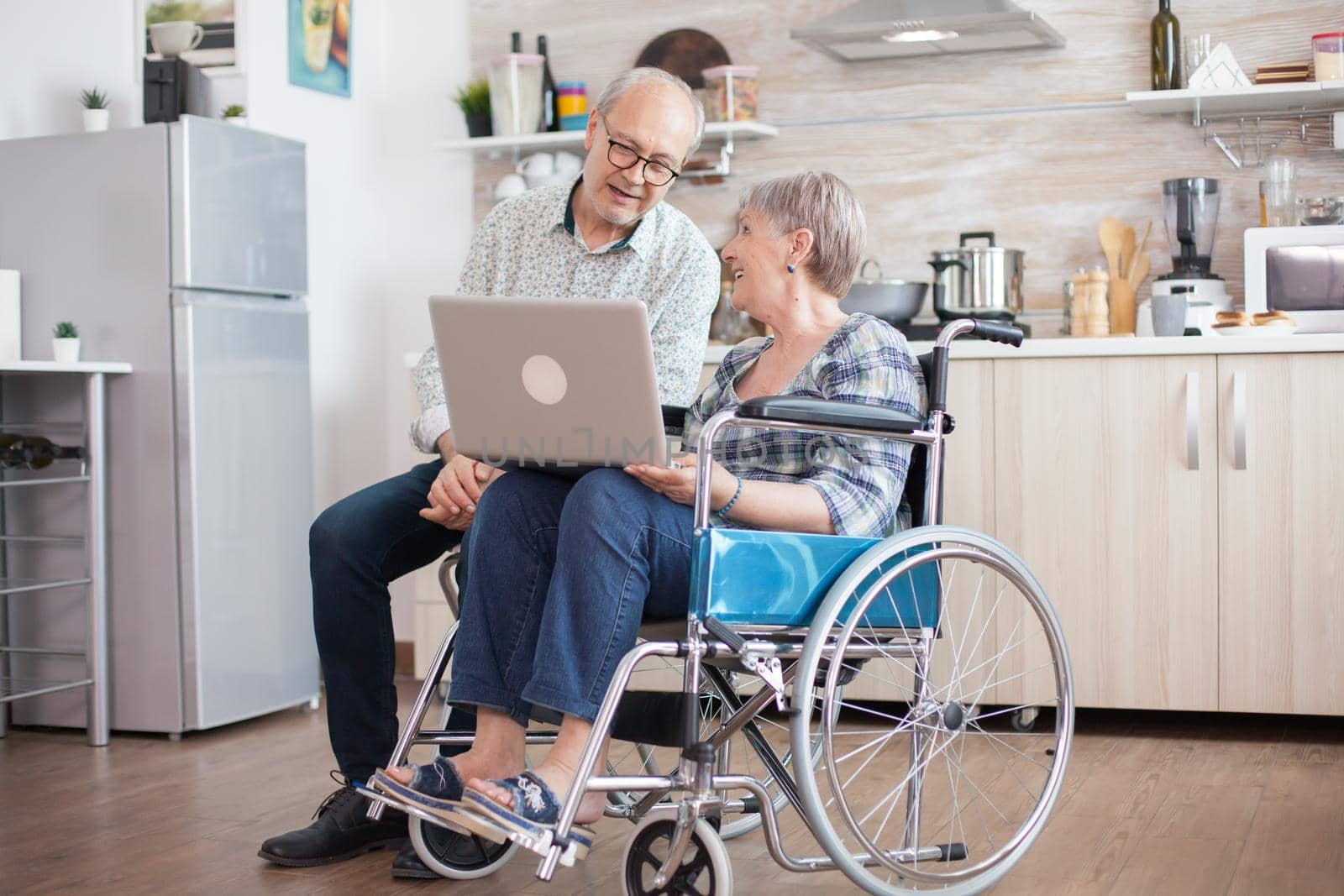 Disabled senior woman in wheelchair and her husband having a video conference on tablet pc in kitchen. Paralyzed old woman and her husband having a online conference.