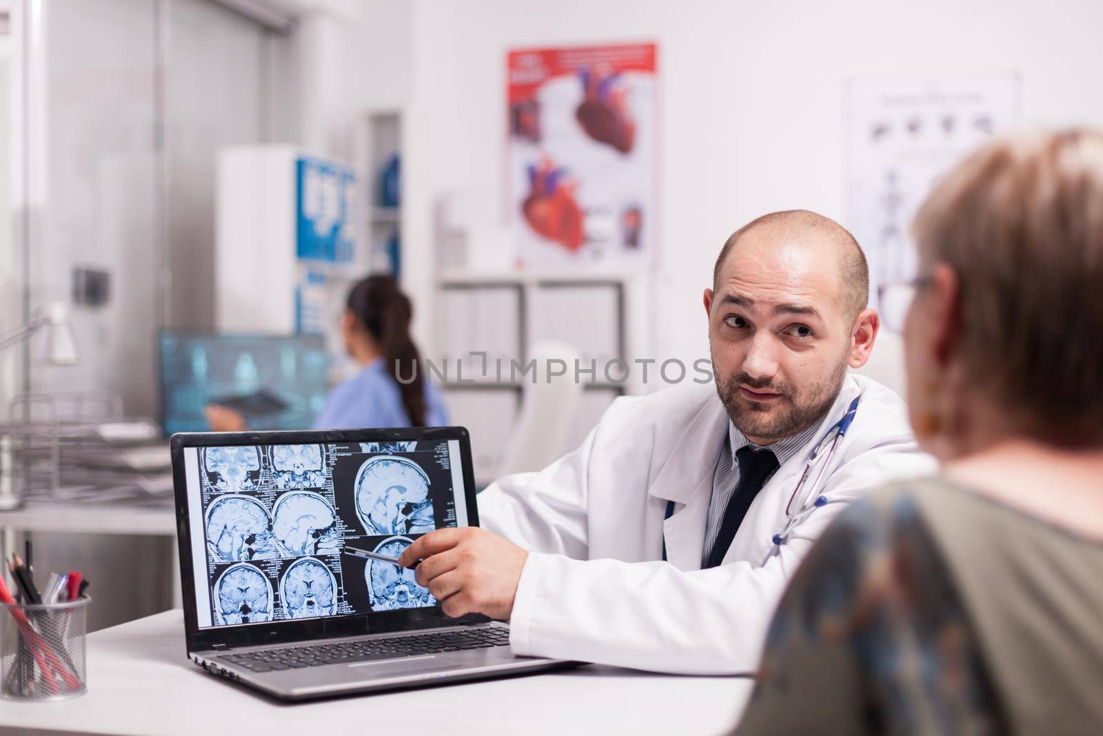 Doctor discussing with senior woman with alzheimer in hospital office pointing at laptop screen with ct scan. Medic wearing white coat. Nurse in blue uniform working on computer.