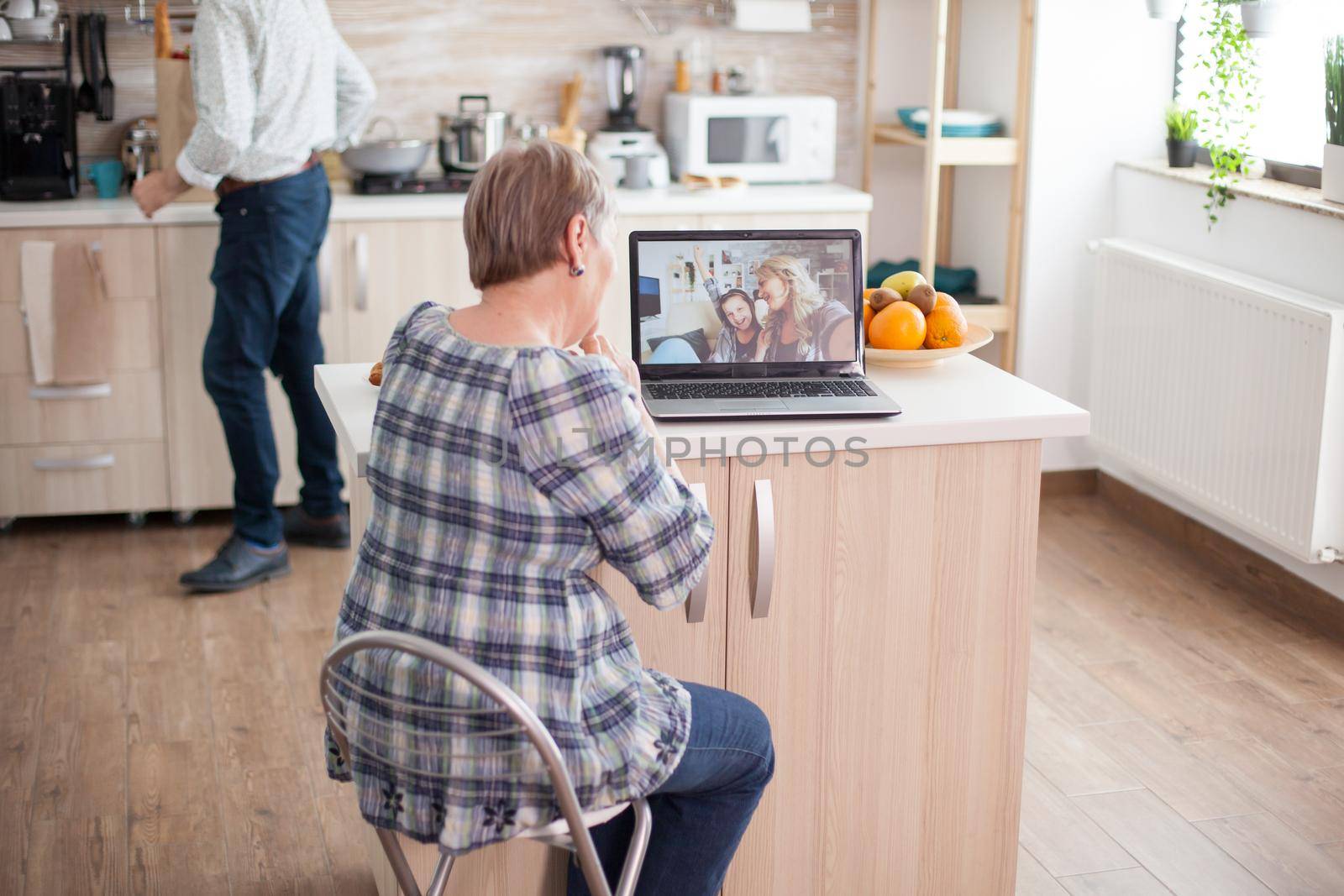 Happy senior woman during a video conference with family using laptop in kitchen. Online call with daughter and niece. Elderly person using modern communication online internet web techonolgy.