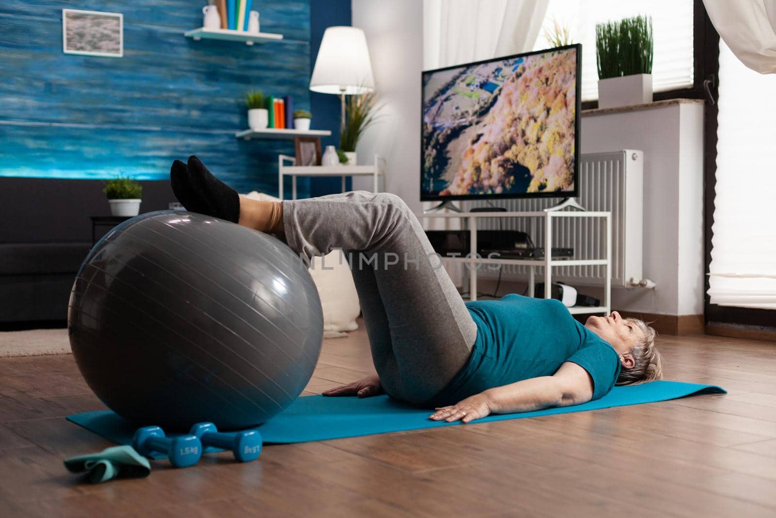 Retired senior woman doing practicing legs up exercise using swiss ball sitting on yoga mat in living room. Retired pensioner in sportswear practicing abs cardio working at body muscular healthcare