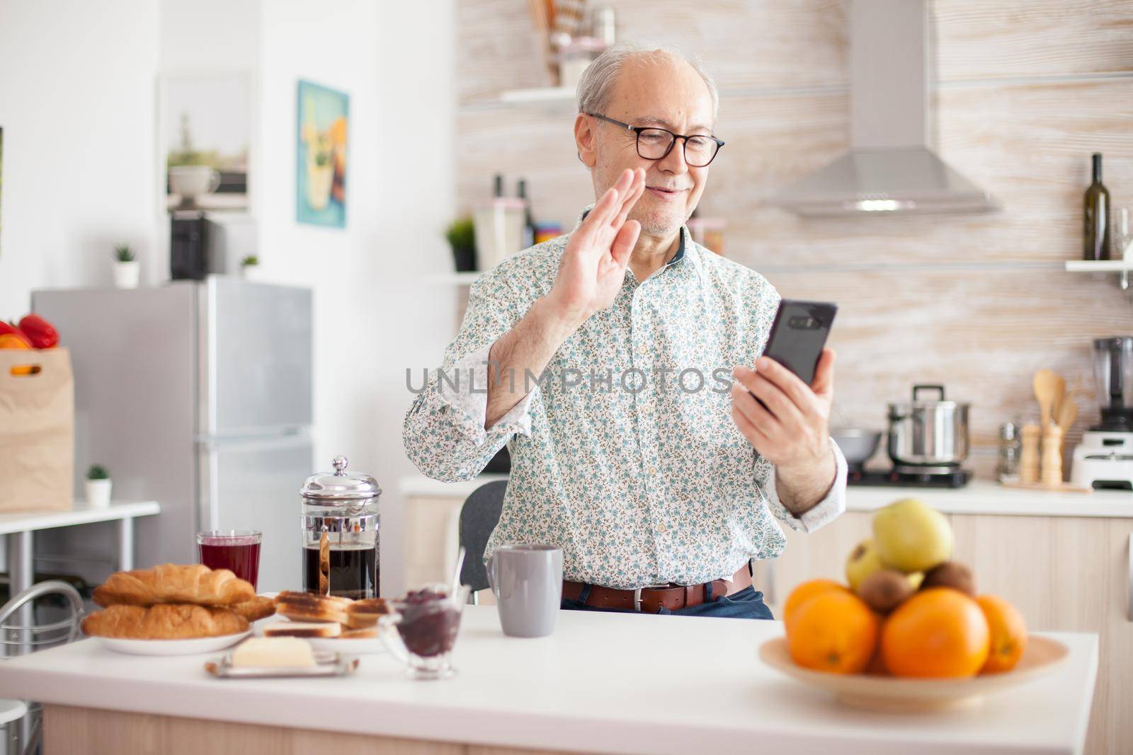 Elderly aged man waving during a video conference with family in kitchen. Elderly person using internet online chat technology video webcam making a video call connection camera communication conference call