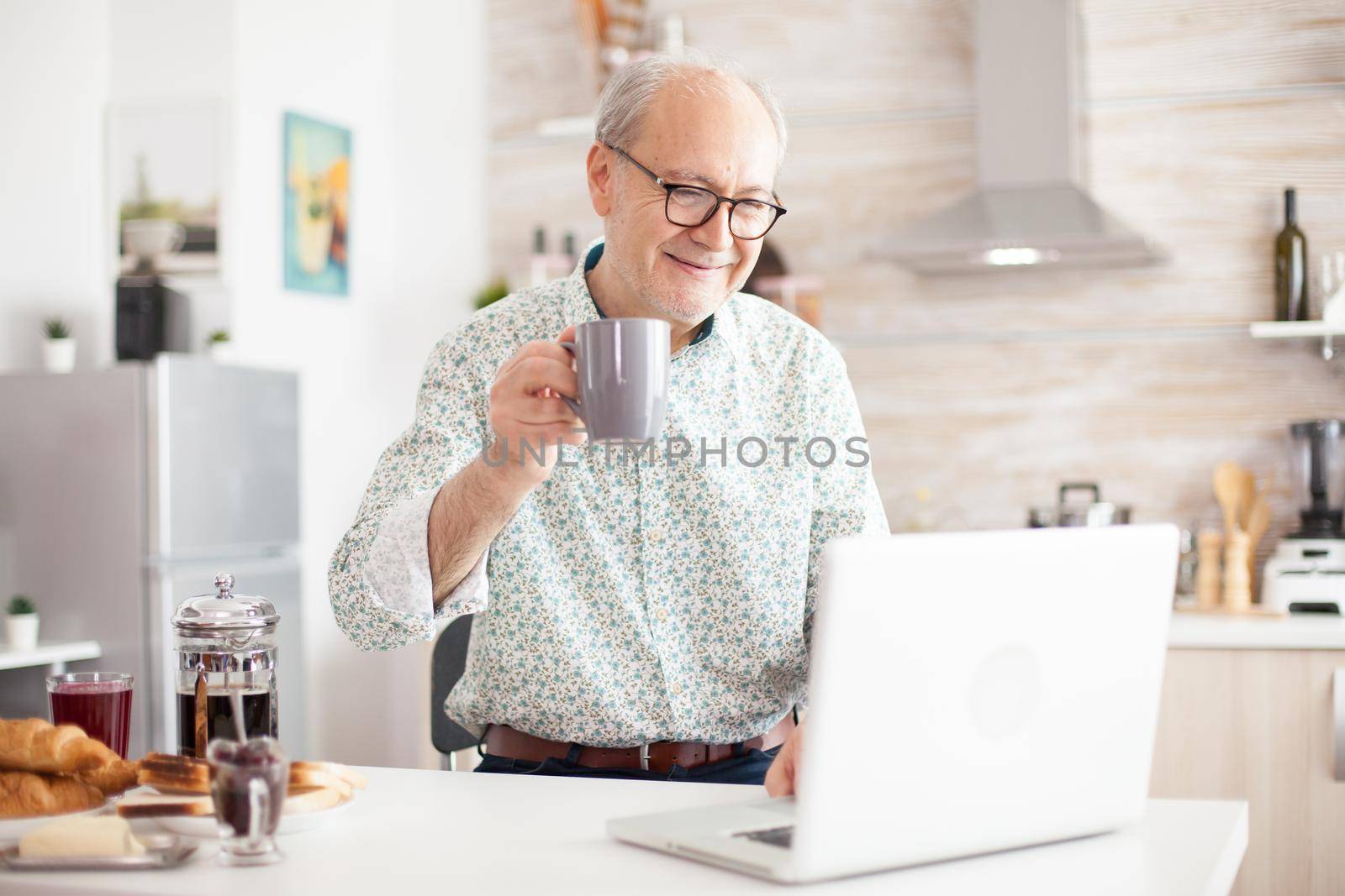 Cheerful senior man during video conference in kitchen while enjoying breakfast and cup of coffee. Elderly person using internet online chat technology video webcam making a video call connection camera communication conference call
