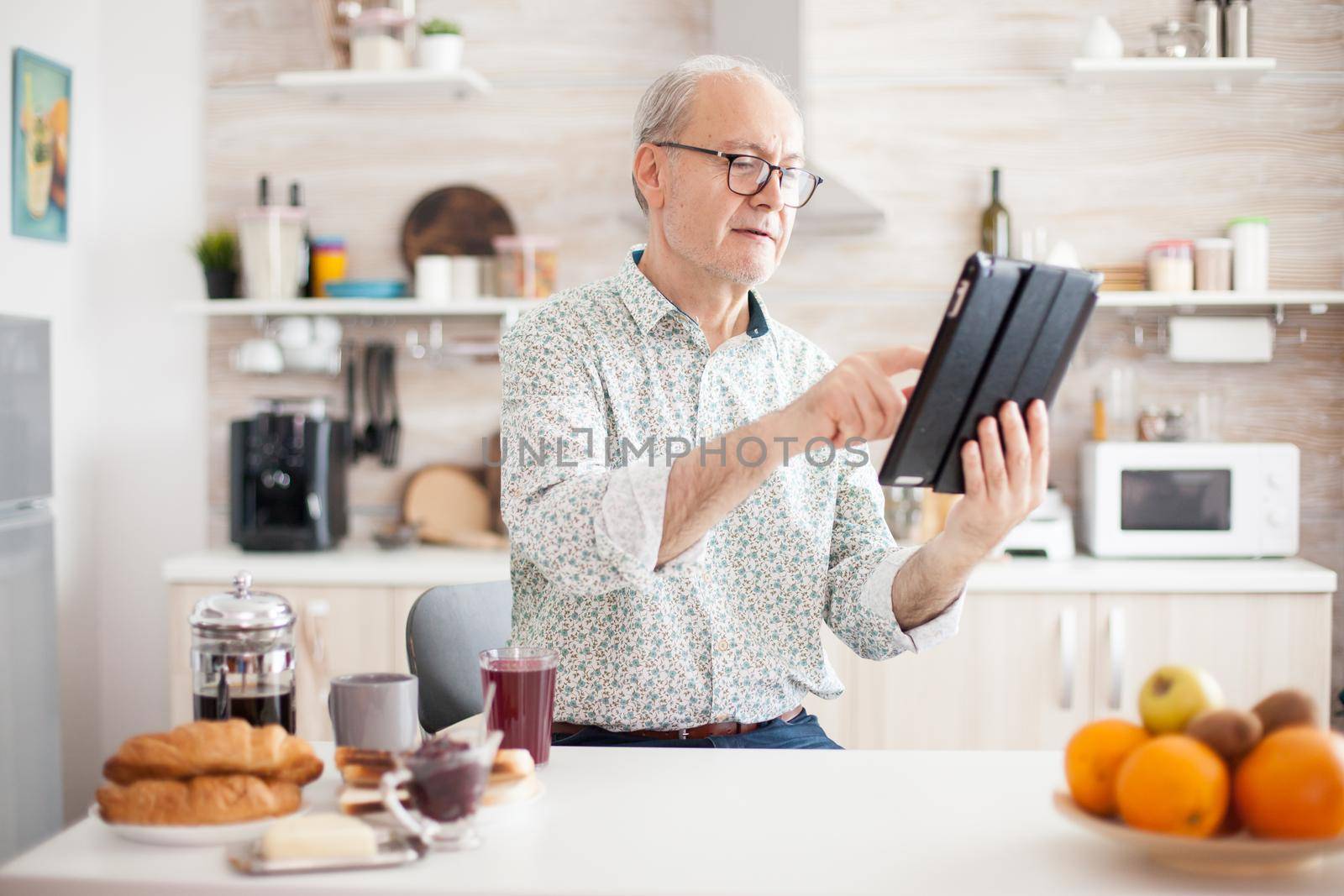 Happy elderly person with portable tablet PC in kitchen during breakfast. Senior person with tablet portable pad PC in retirement age using mobile apps, modern internet online information technology with touchscreen