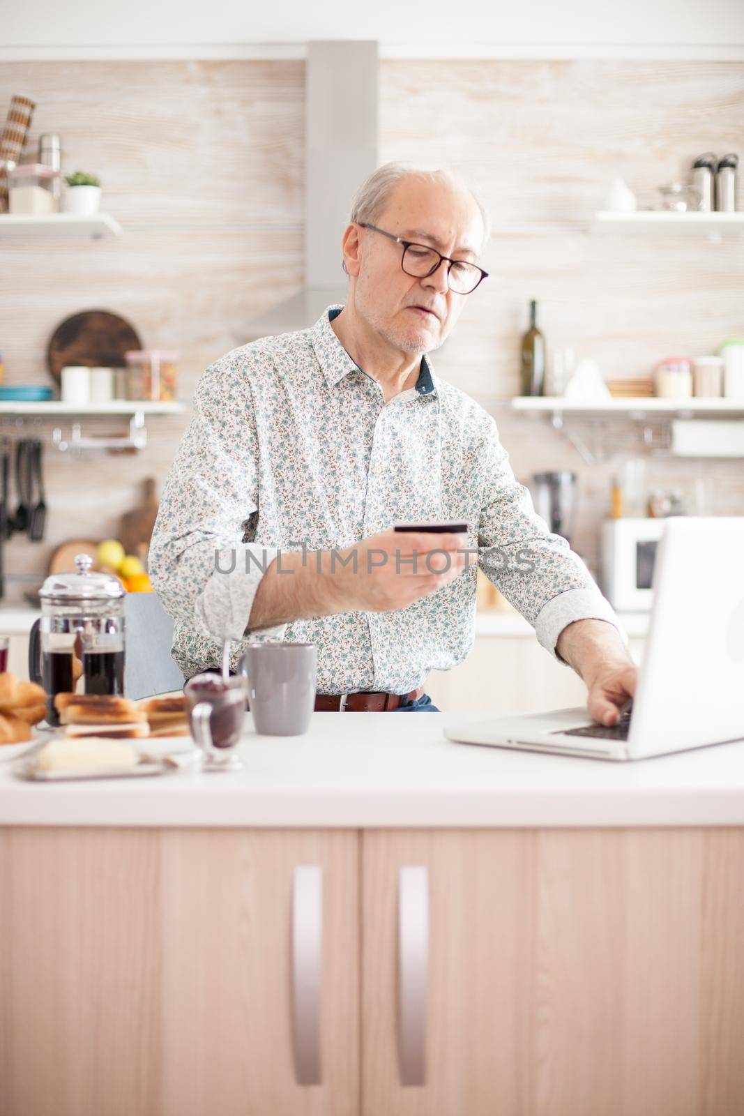 Elderly man checking finances, holding a credit card in his hand. Pensioner paying online using credit card and application from laptop during breakfast in kitchen. Retired elderly person using internet payment home bank buying with modern technology