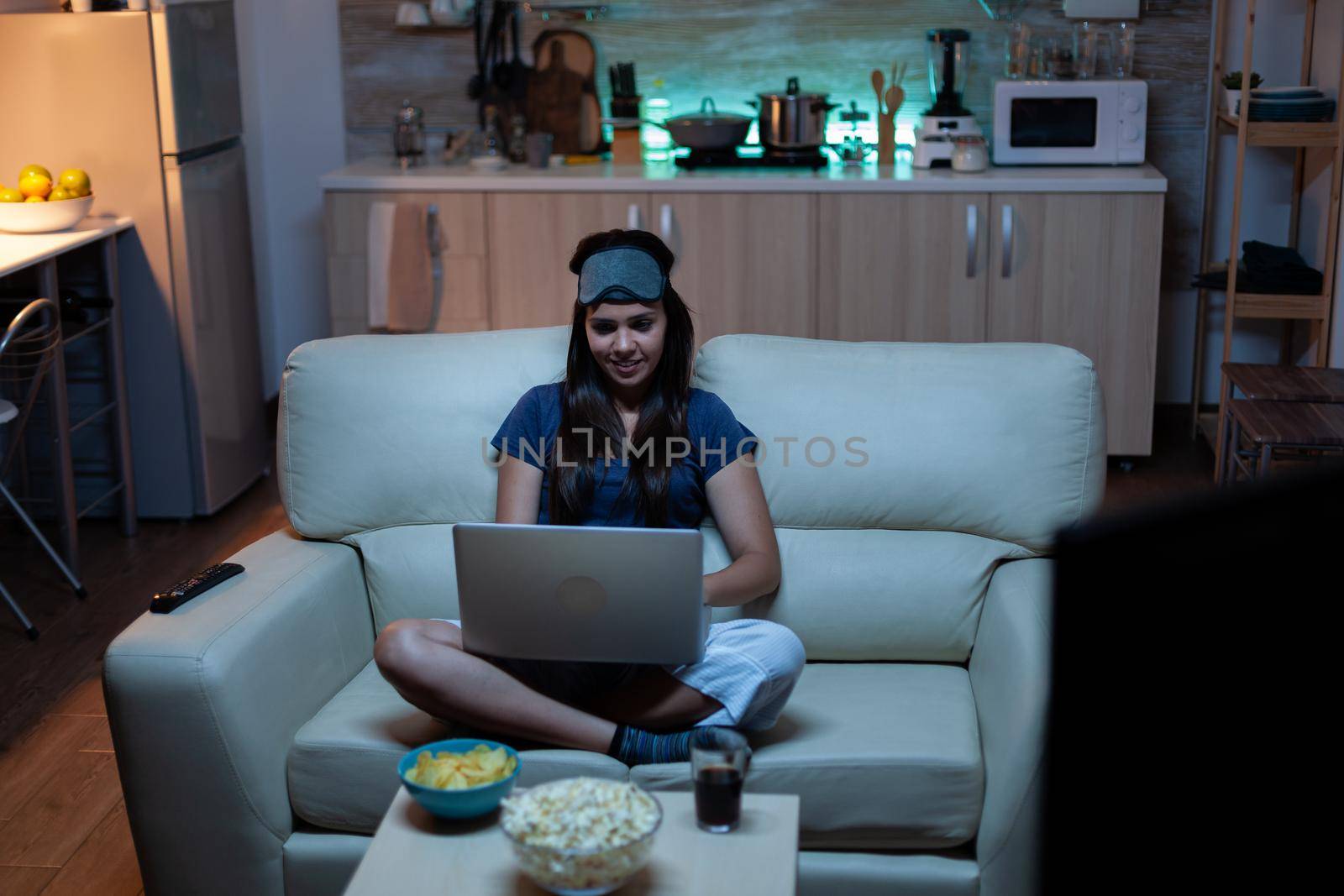 Lady in pijamas and eye cover on forehead typing on laptop and watching tv late at night . Freelancer working sitting on sofa reading writing searching browsing on notebook using internet technology