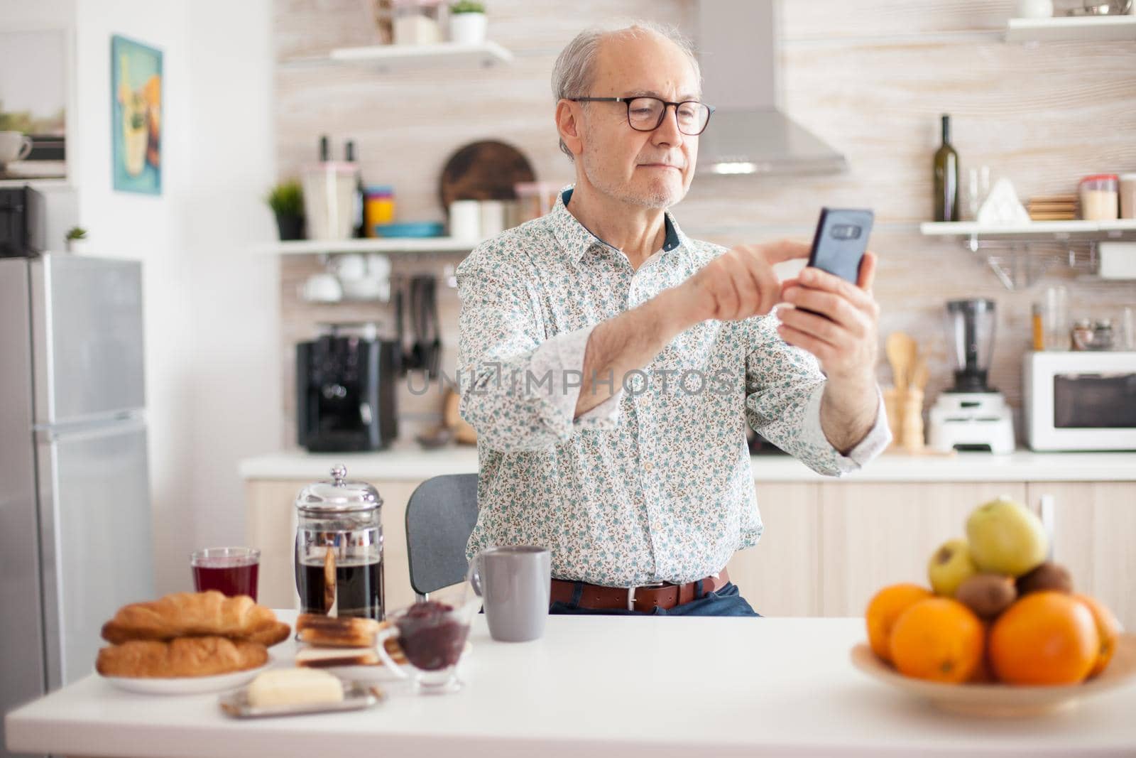 Happy senior man browsing the internet on smartphone during breakfast in kitchen. Elderly person using internet online chat technology video webcam making a video call connection camera communication conference call