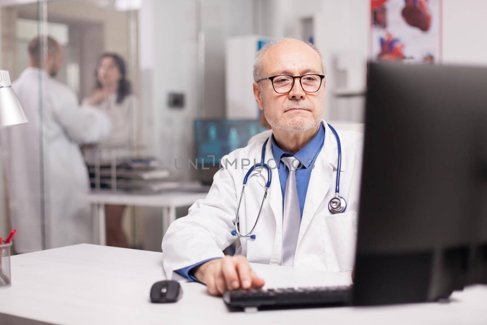 Senior doctor typing expertise rapport on computer in clinic office while young therapist in white coat assisting patient on hospital corridor.
