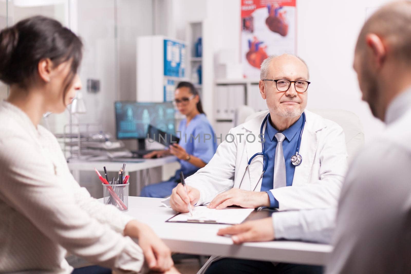 Senior medical practitioner taking notes on clipboard during consultation of married couple in hospital office. Doctor looking at male patient while wearing white coat and stethoscope.
