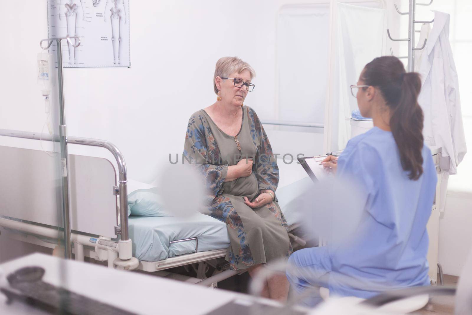 Pensioner woman during medical check with nurse in hospital office. Assistant in blue uniform taking notes on clipboard.