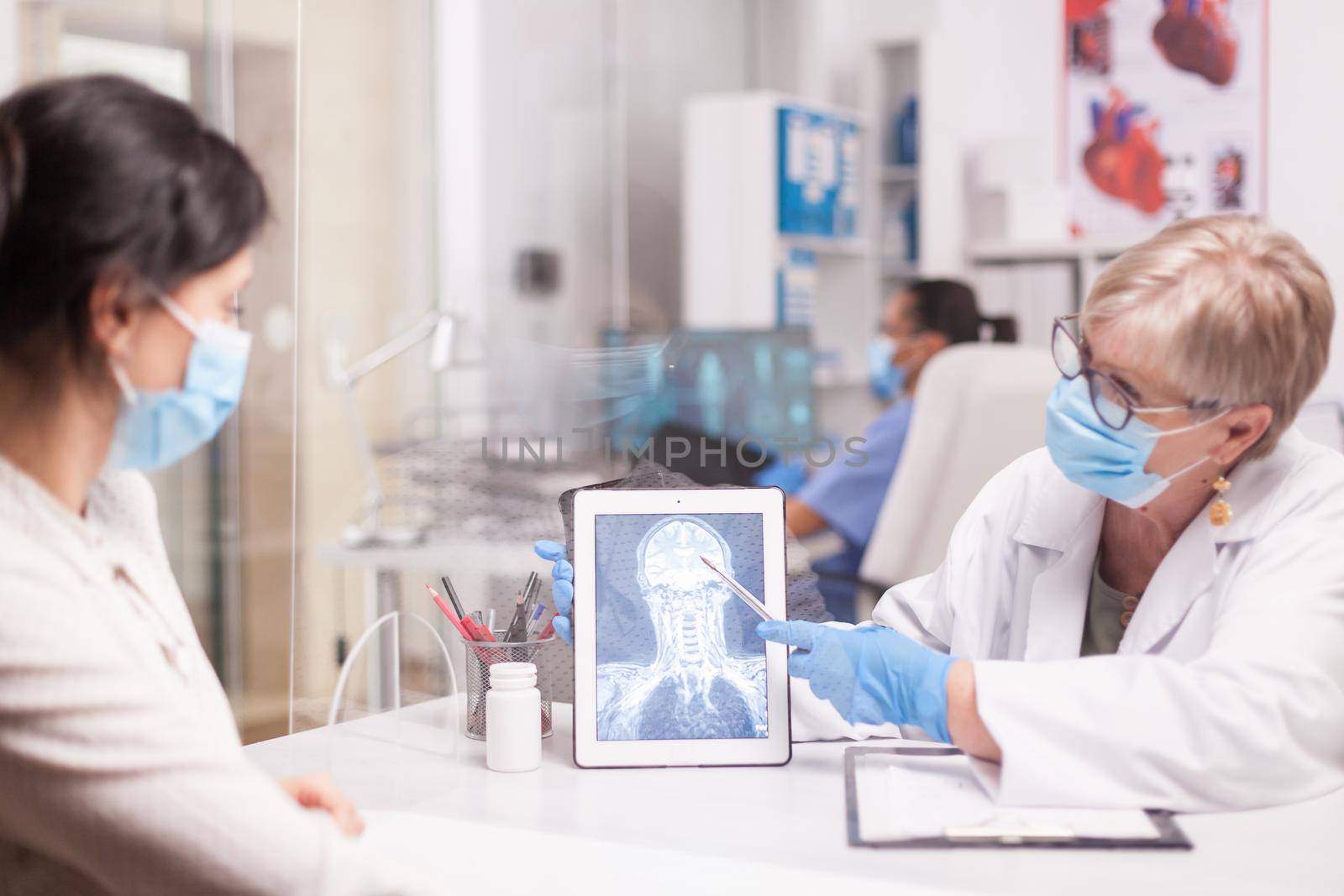 Senior doctor with face mask against covid-19 pointing at brain trauma on table computer during patient examination. Young woman with skull injury.