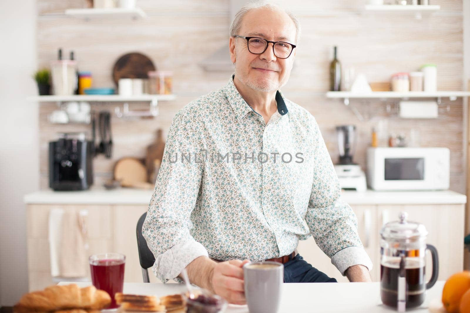 Senior man in kitchen smiling looking at camera holding hot coffee cup. Portrait of relaxed elderly older person in the morning, enjoying fresh warm drink. Healthy smiling adult face