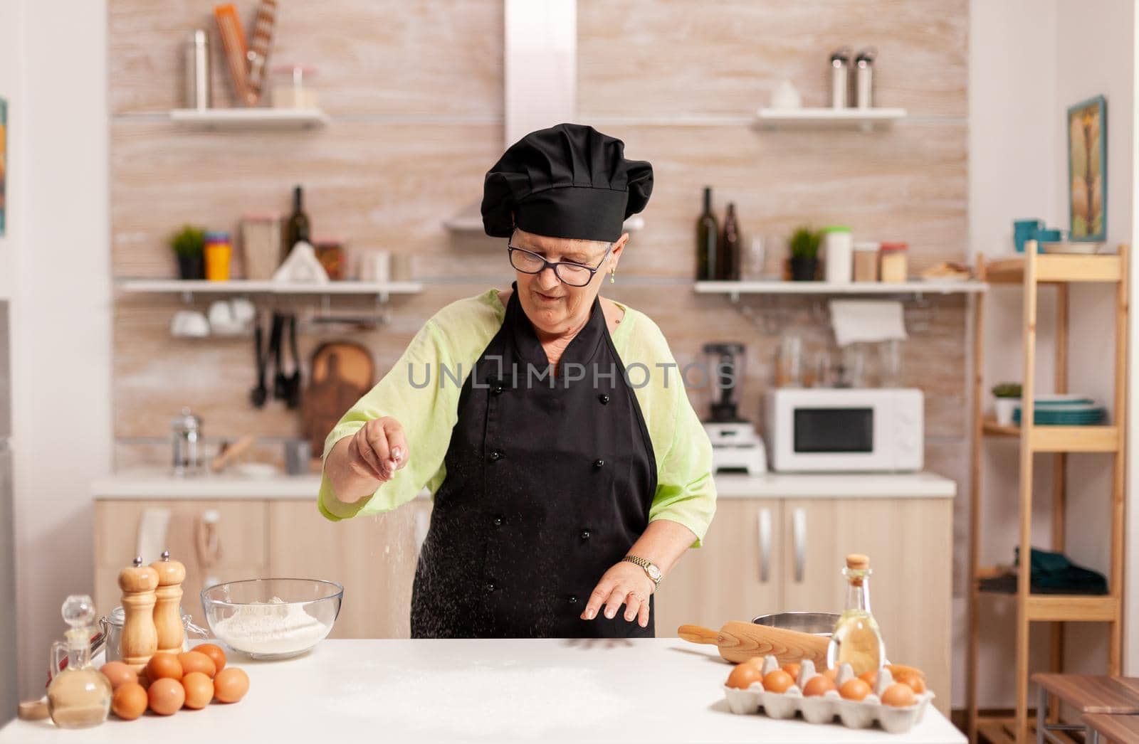 Woman preparing homemade cookies spreading flour table in home kitchen. Happy elderly chef with uniform sprinkling, sieving sifting raw ingredients by hand baking homemade pizza