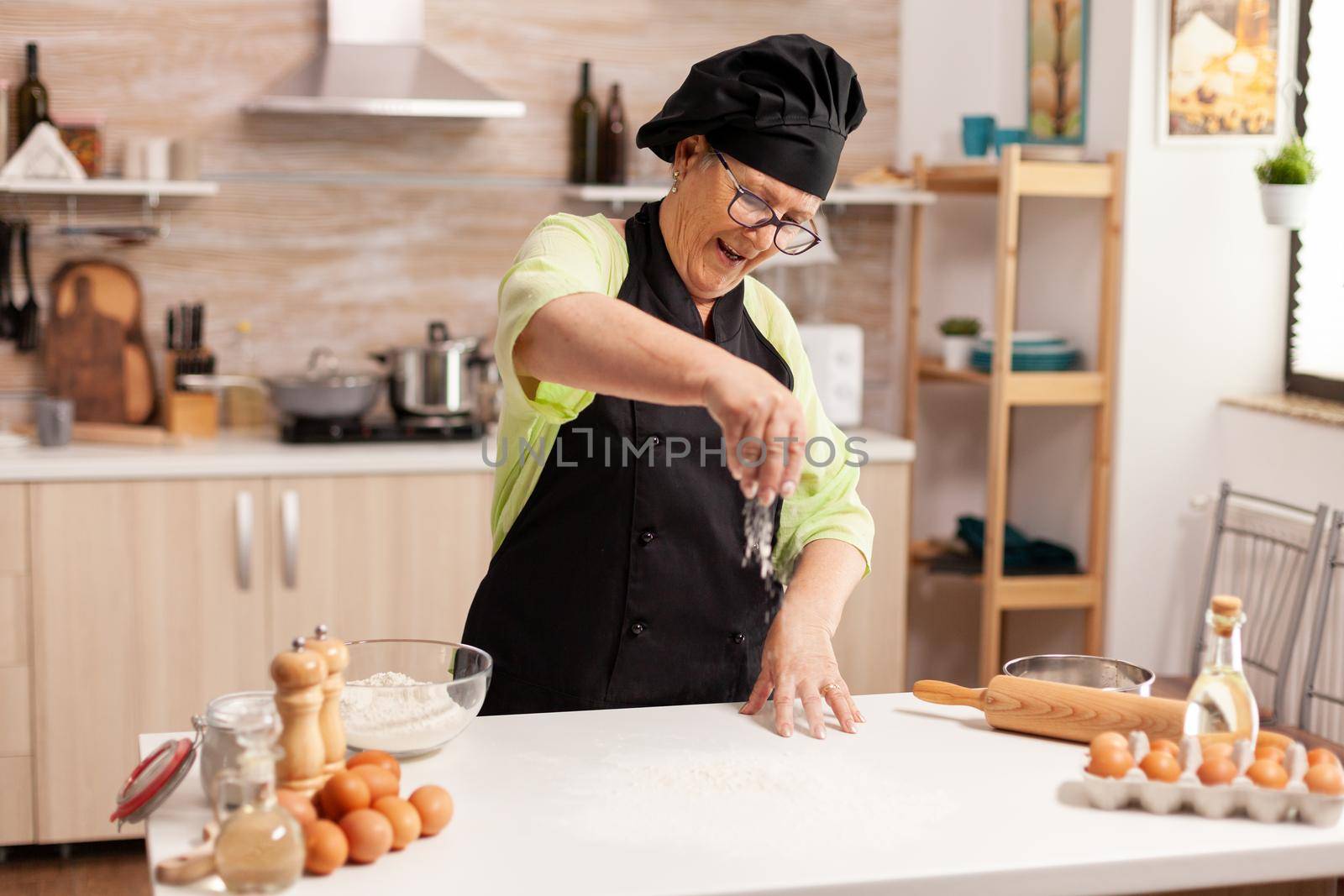 Cheerful senior lady making pizza in home kitchen using top flour for baking. Happy elderly chef with uniform sprinkling, sieving sifting raw ingredients by hand.