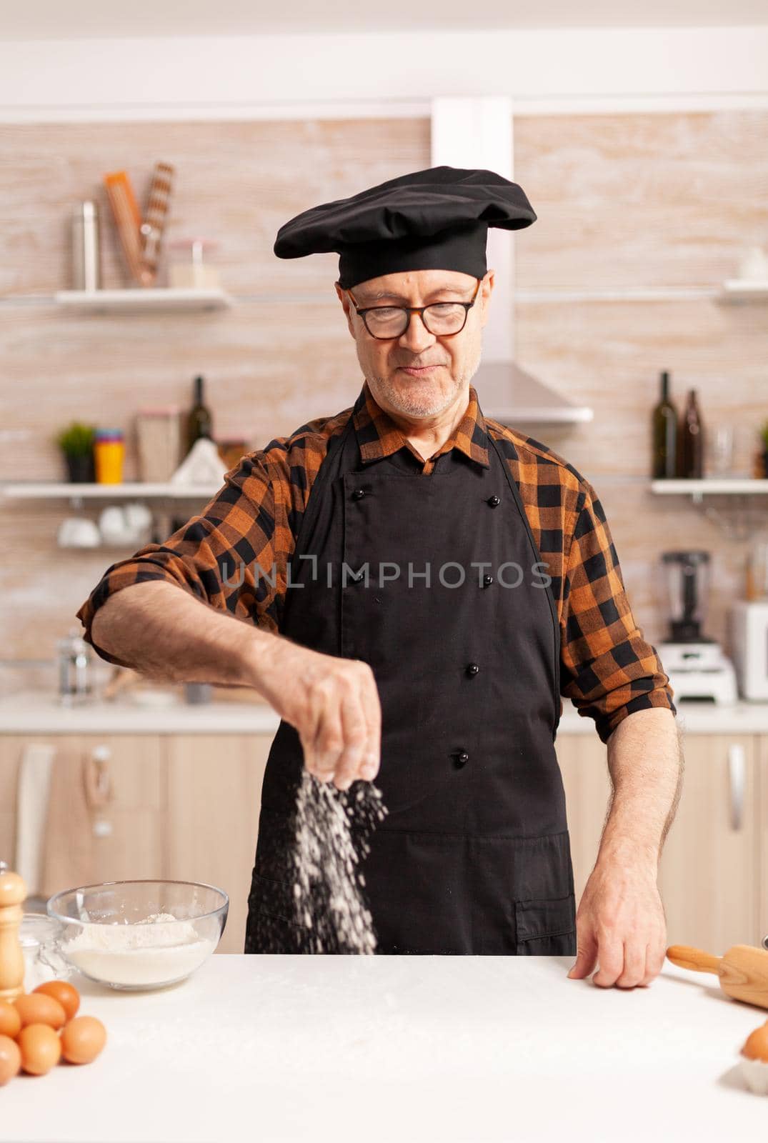Happy retired chef preparing homemade pizza using bio wheat flour. Retired senior chef with bonete and apron, in kitchen uniform sprinkling sieving sifting ingredients by hand.