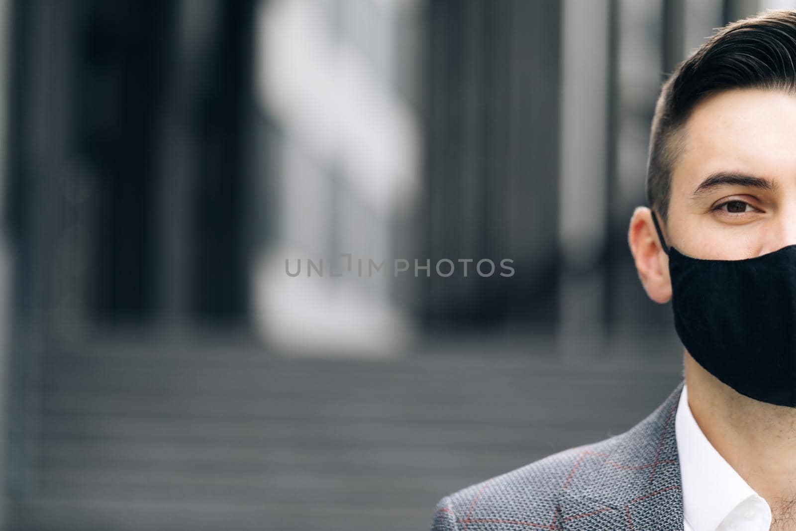 Half face of upset caucasian young handsome man in medical mask looking straight to camera while standing outdoors near modern office building. Man in protection mask look at camera.