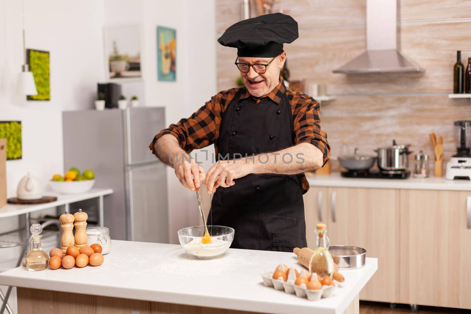 Retired chef cracking eggs for wheat flour in home kitchen. Elderly pastry chef cracking egg on glass bowl for cake recipe in kitchen, mixing by hand, kneading.