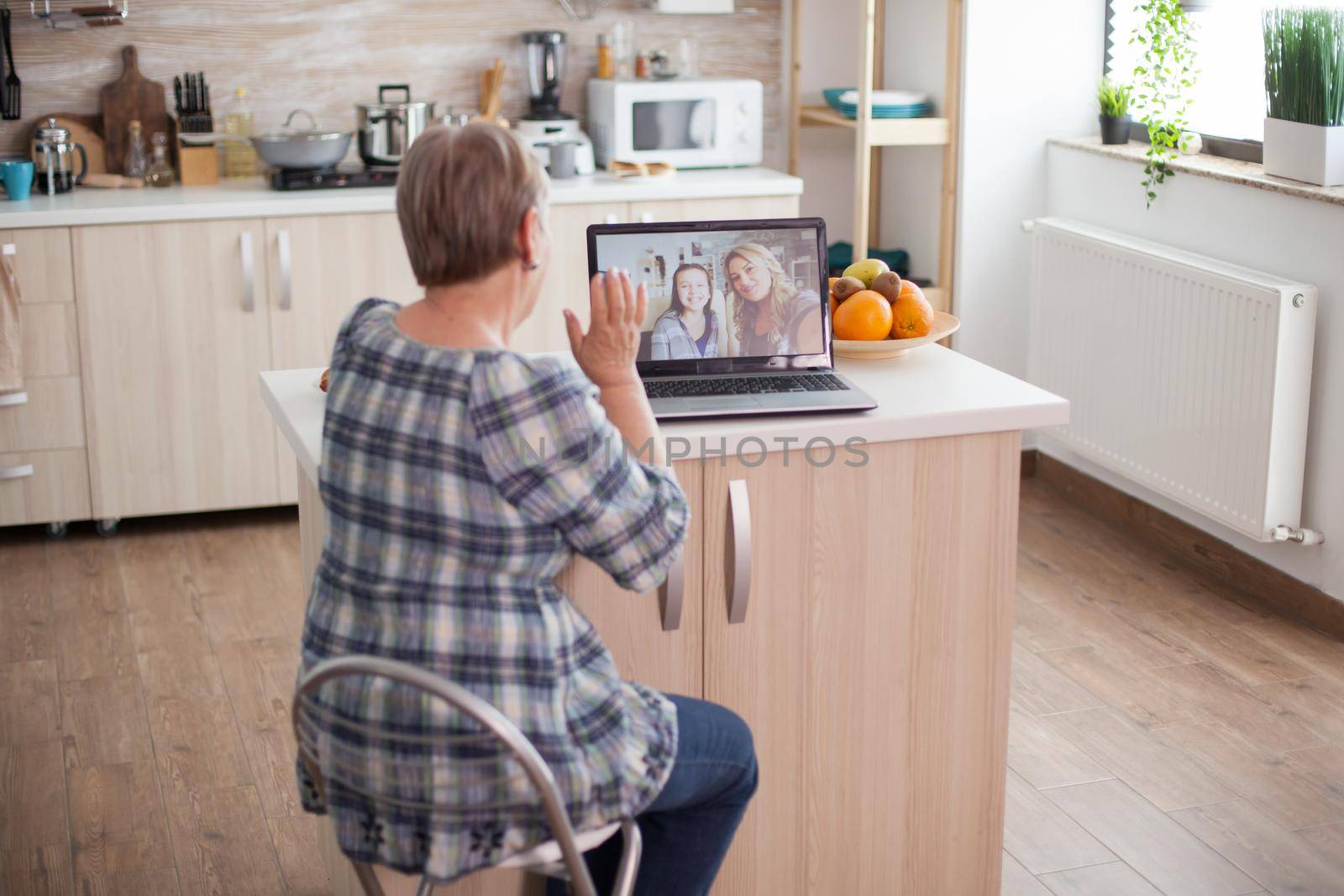 Senior woman waving to her daughter. Happy senior woman during a video conference with family using laptop in kitchen. Online call with daughter and niece. Elderly person using modern communication online internet web techonolgy.