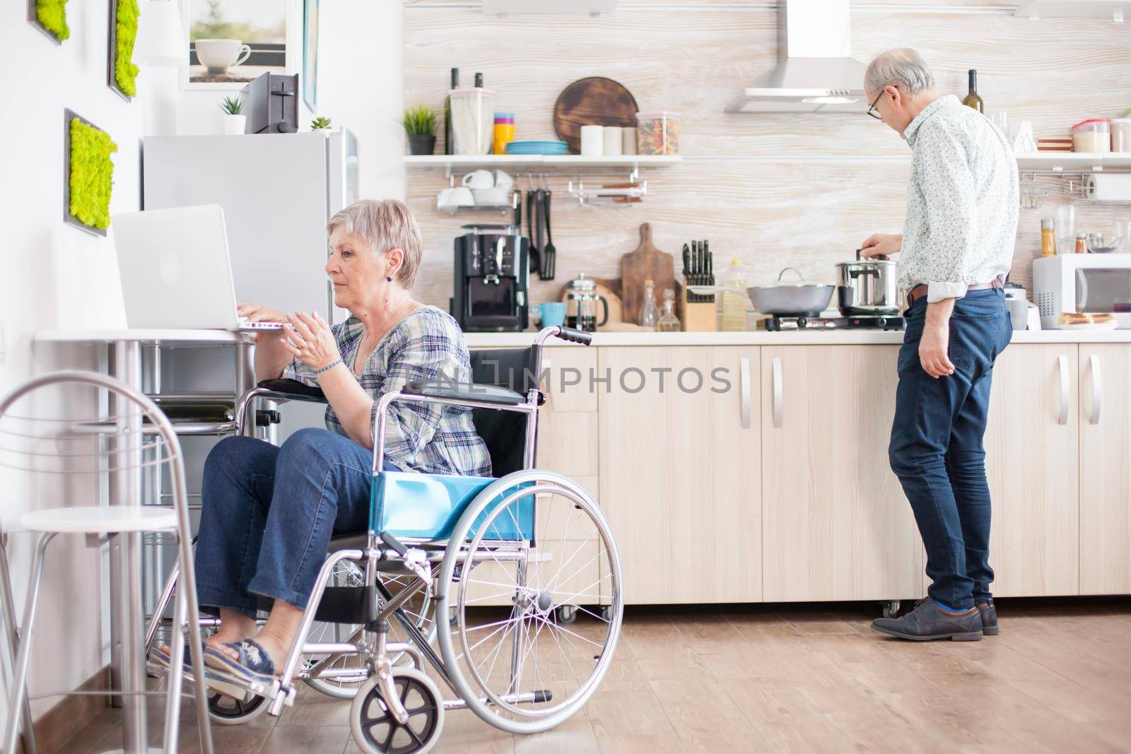 Disabled old woman in wheelchair working on laptop in kitchen. Paralyzed handicapped old elderly person using modern communication online internet web techonolgy.