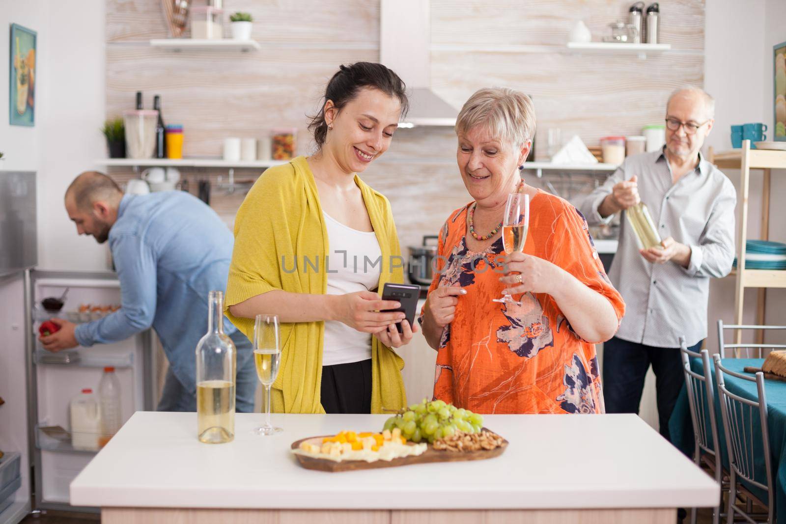 Mother and daughter surfing internet using smarthphone in kitchen during family meeting and drinking wine.