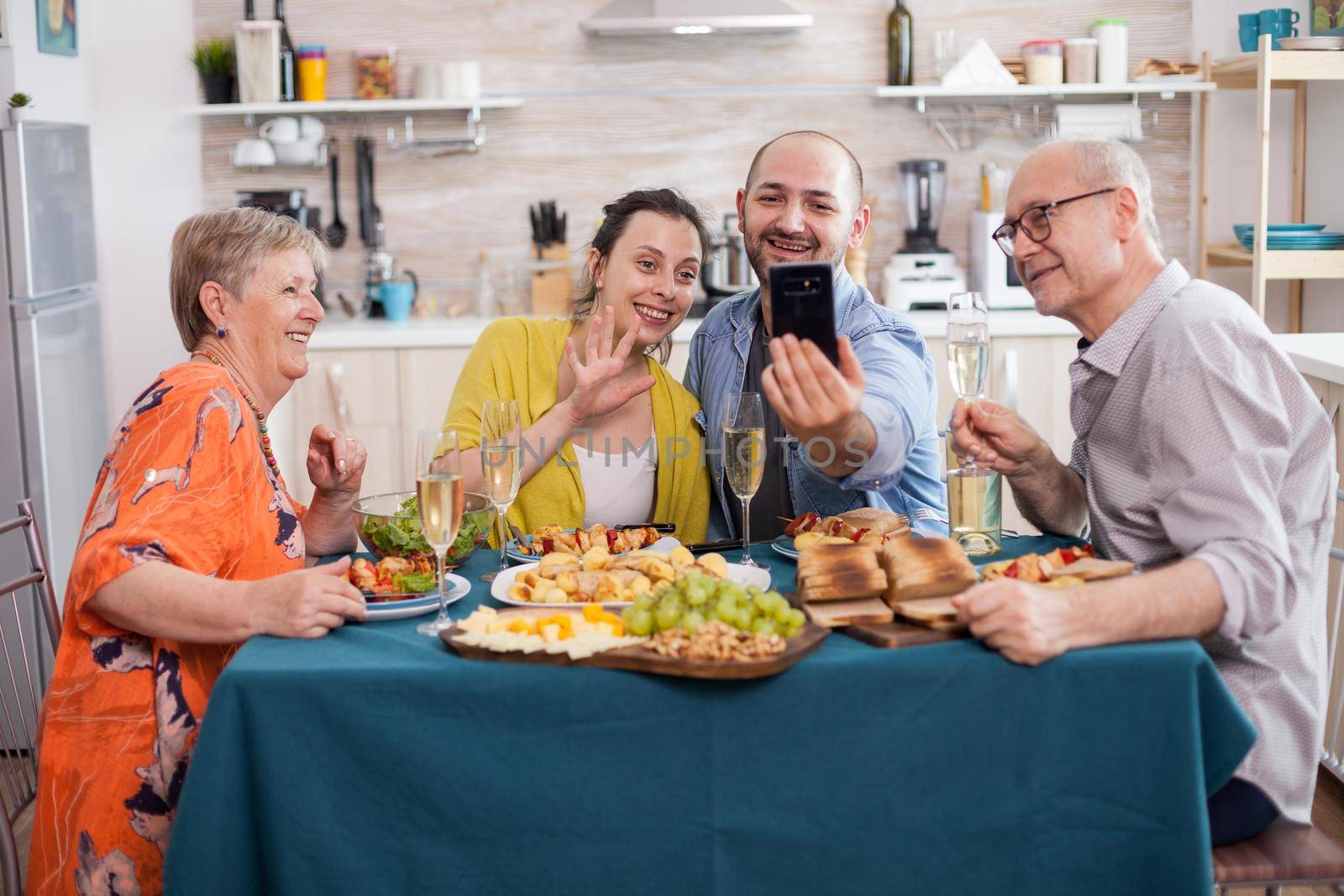 Cheerful family during video call while having delicious brunch in kitchen at dining table. Parents bonding with son and his wife.