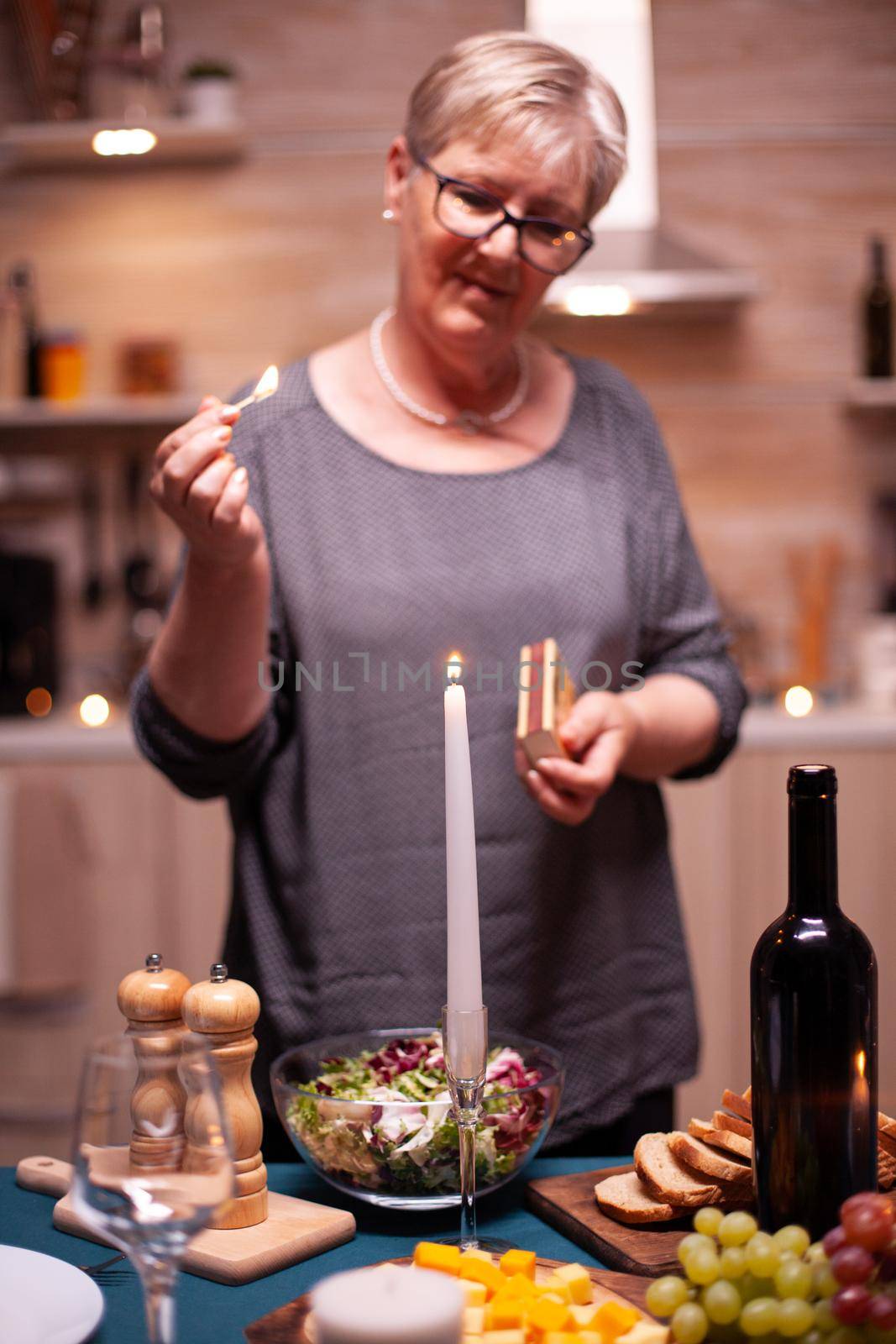 Wife looking at burning candle while preparing table for romantic dinner with husband. Elderly woman waiting her husband for a romantic dinner. Mature wife preparing festive meal for anniversary celebration.