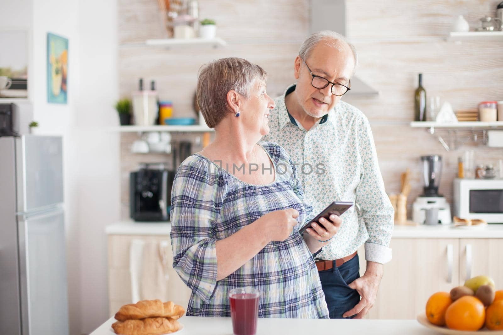 Senior woman holding smartphone and sharing the recipe with husband. Caucasian senior wife and husband in kitchen during breakfast using smartphone technology and internet conection. Leisure retired woman and man smiling in domicile, relaxing.