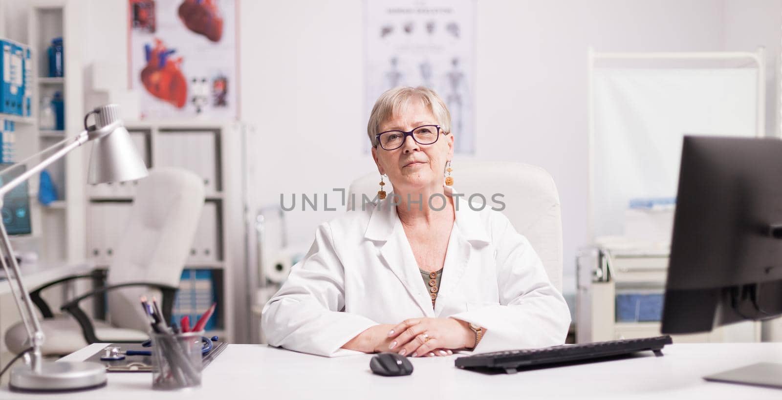 Elderly aged woman doctor in hospital cabinet wearing white coat.