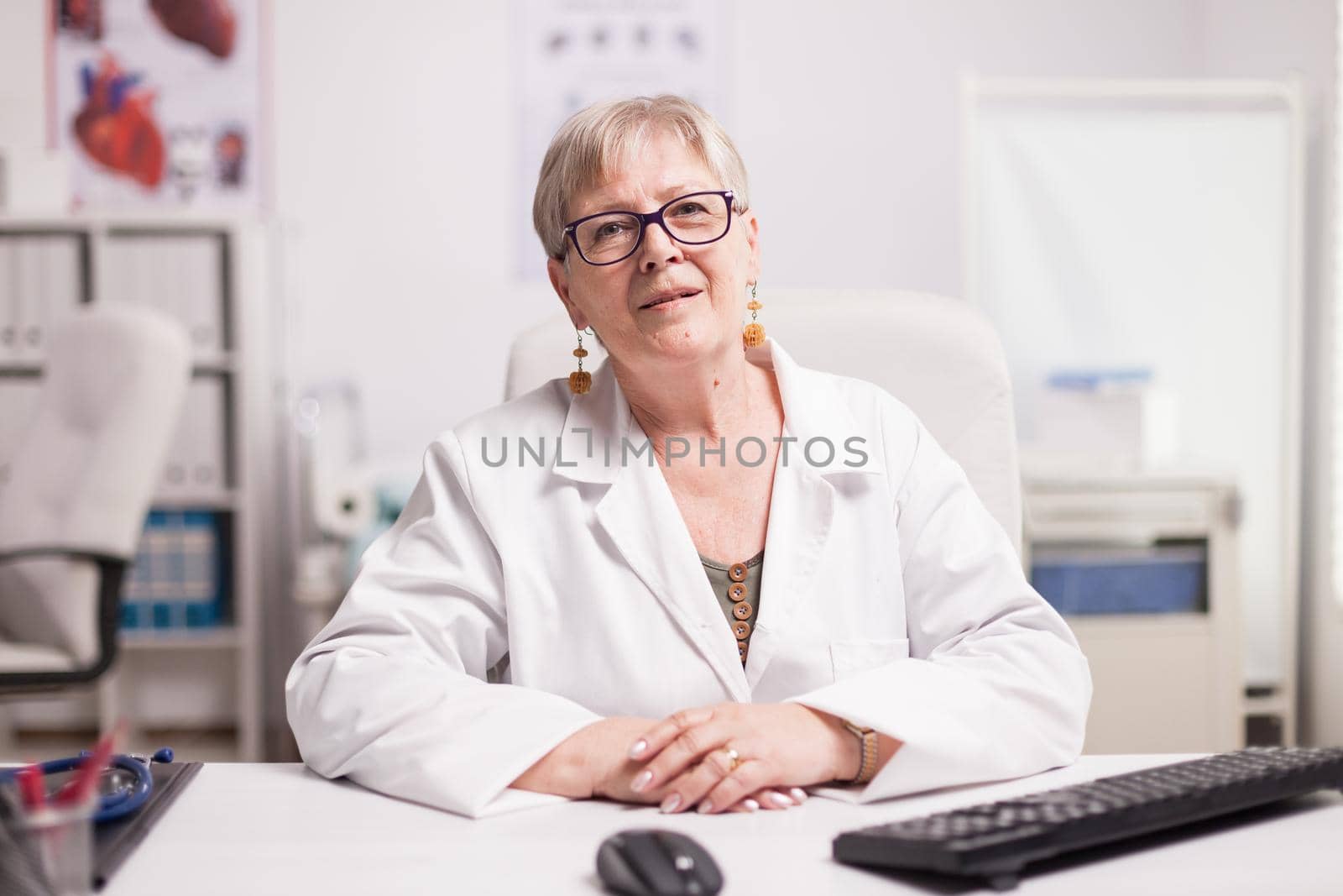Medic with grey hair wearing coat in clinic cabinet sitting at desk.