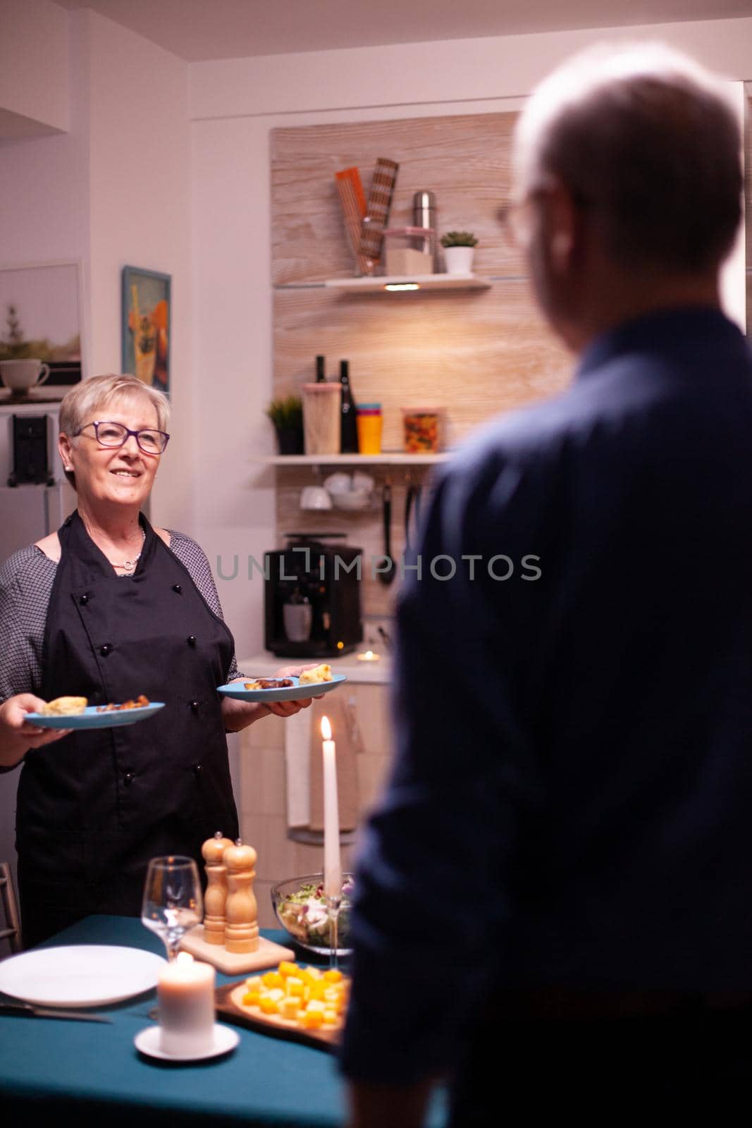 Romantic senior wife in kitchen talking with husband while serving romantic dinner. Elderly old couple talking, sitting at the table in kitchen, enjoying the meal, celebrating their anniversary .