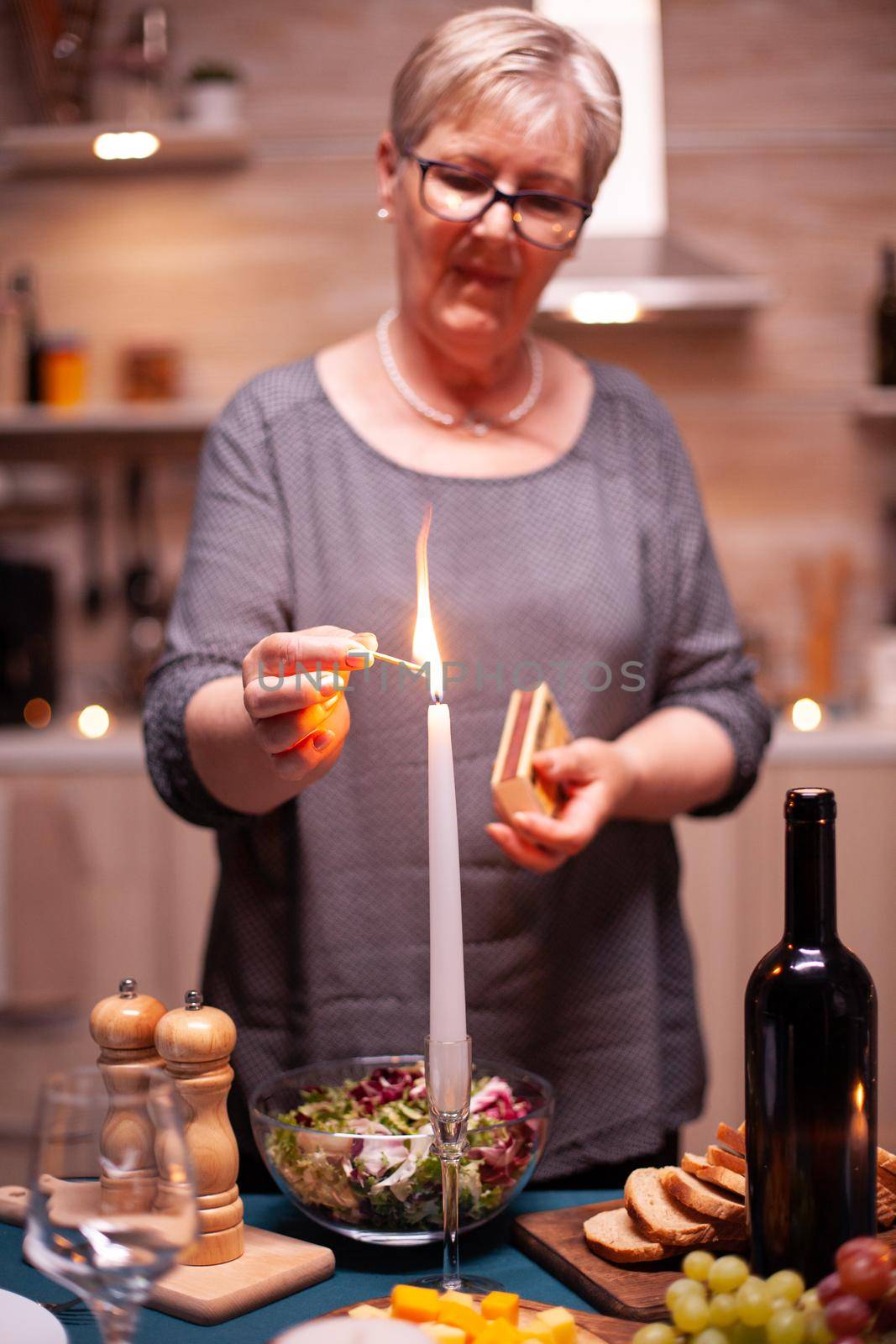 Candle burning on kitchen table for romantic dinner. Elderly woman waiting her husband for a romantic dinner. Mature wife preparing festive meal for anniversary celebration.