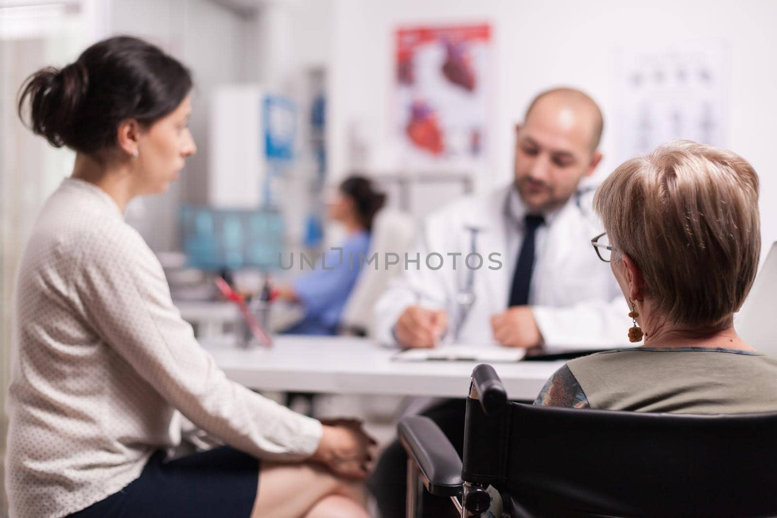 Disabled old woman in wheelchair with daughter listening doctor during consultation in hospital office. Medic wearing white coat and stethoscope taking notes during examination.