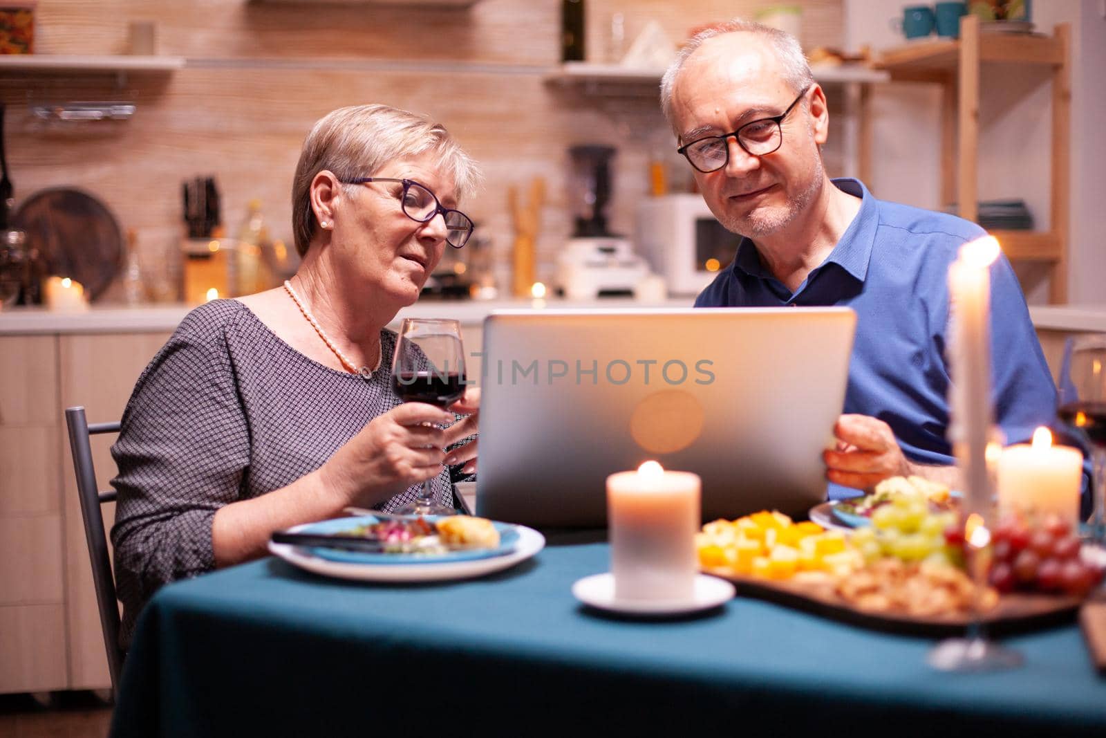 Aged man using laptop in kitchen during relationship celebration with wife. Elderly people sitting at the table browsing, searching, using laptop, technology, internet, celebrating their anniversary in the dining room.