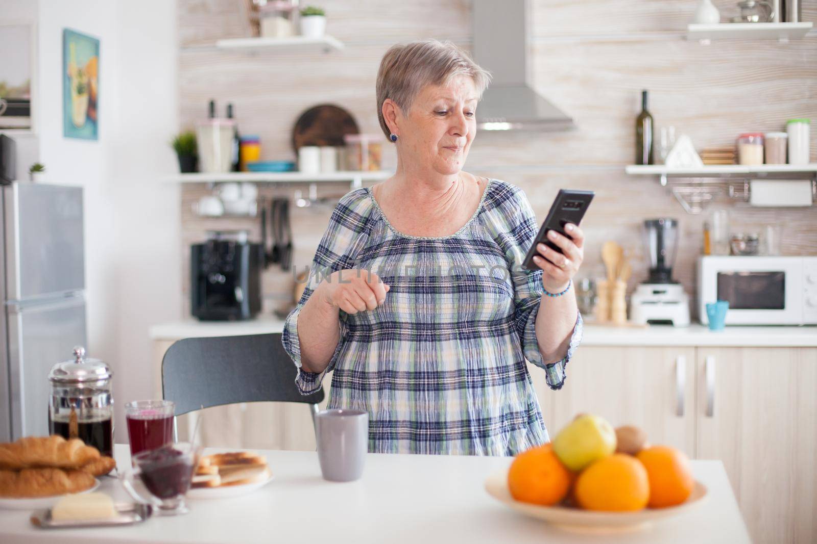 Old woman enjoying music from smartphone during breakfast in kitchen holding cup of coffee. Relaxed elderly dancing, fun lifestyle with modern technology