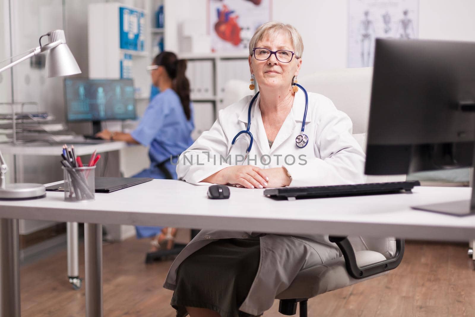 Mature medical therapist sitting at desk in hospital cabinet while nurse in blue uniform working on computer.