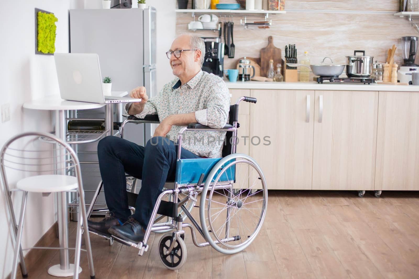 Senior man in wheelchair using laptop in the kitchen. Disabled senior man in wheelchair having a video conference on laptop in kitchen. Paralyzed old man and his wife having a online conference.