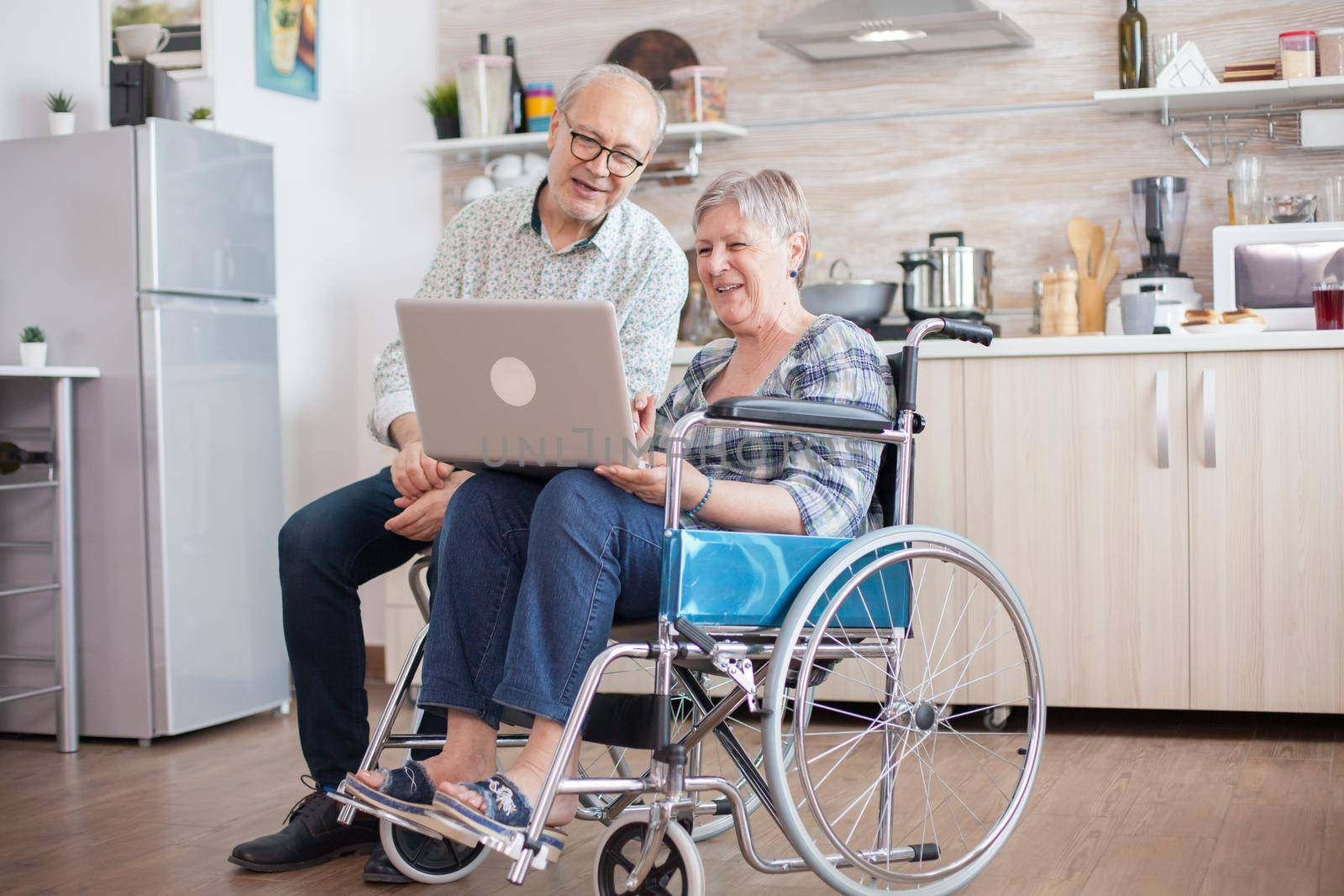 Senior couple looking at webcam before a video call. Disabled senior woman in wheelchair and her husband having a video conference on tablet pc in kitchen. Paralyzed old woman and her husband having a online conference.