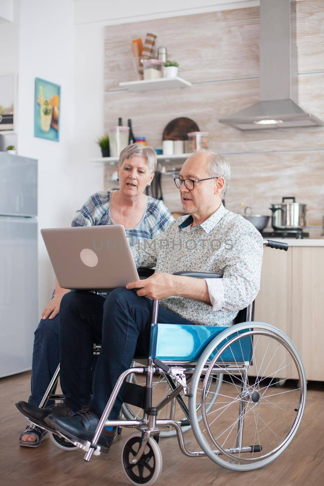 Disabled senior man in wheelchair and his wife are browsing on internet using laptop . Paralysed man using modern technology. Wife talking with husband.