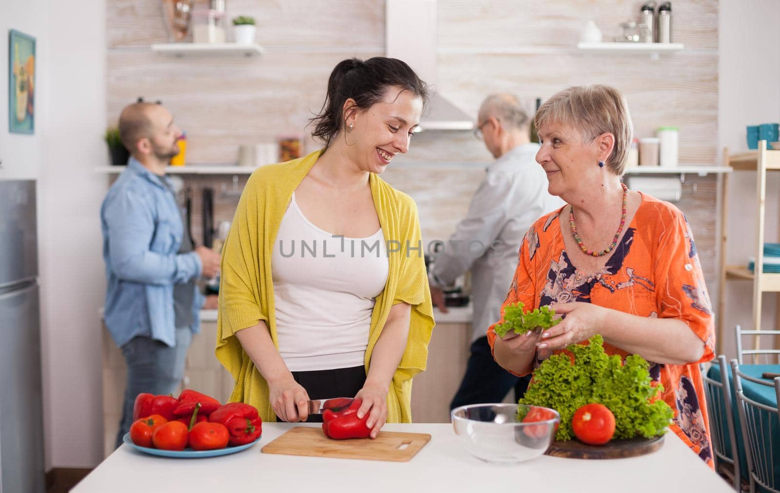 Mother and daughter looking at each other while making salad from fresh vegetables. Husband cooking.