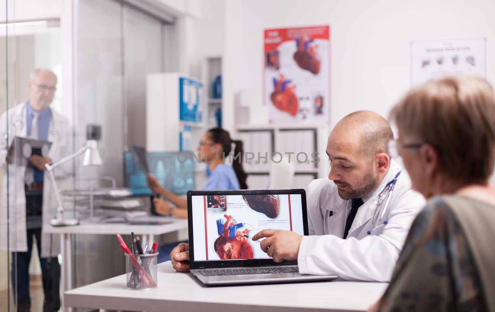 Young doctor pointing at heart on laptop screen in hospital office. Cardiologist with senior patient during consultation. Mature medic taking notes on clipboard on clinic corridor.