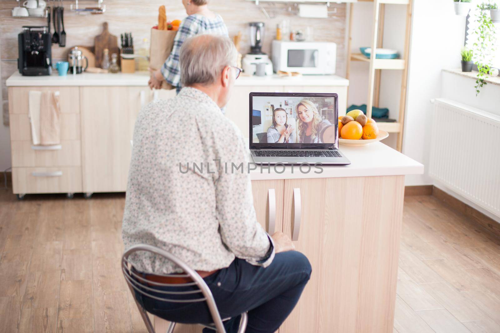 Senior man talking with niece on video call in the kitchen. Happy senior man during a video conference with family using laptop in kitchen. Online call with daughter and niece. Elderly person using modern communication online internet web techonolgy.