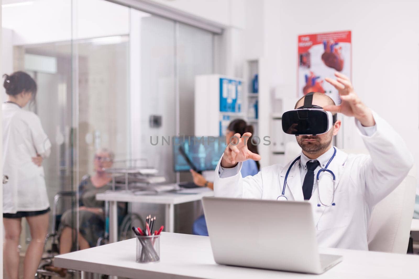 Young physician using virtual reality goggles in hospital office. Invalid senior woman in wheelchair talking with female doctor on clinic corridor. Nurse in blue uniform holding patient x-ray.