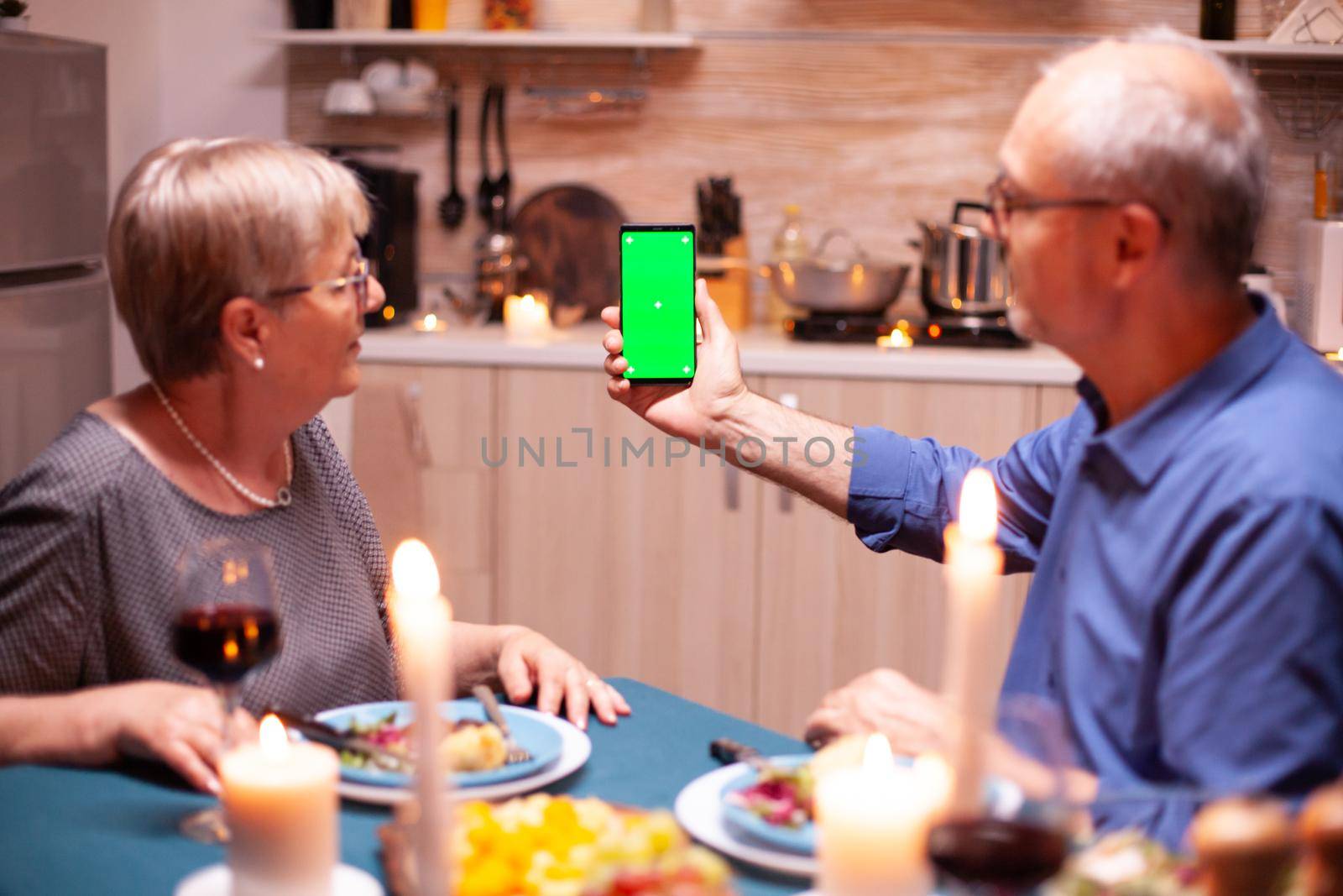 Mature man using phone with green screen in kitchen during romantic dinner with wife. Aged people looking at mockup template chroma key isolated smart phone display using techology internet.